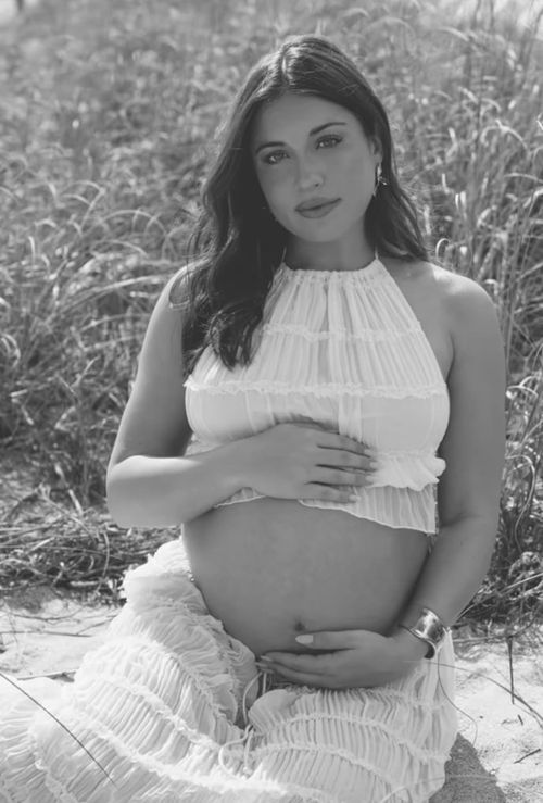 Black and white maternity photoshoot in a field shows a woman in a white dress cradling her baby bump.