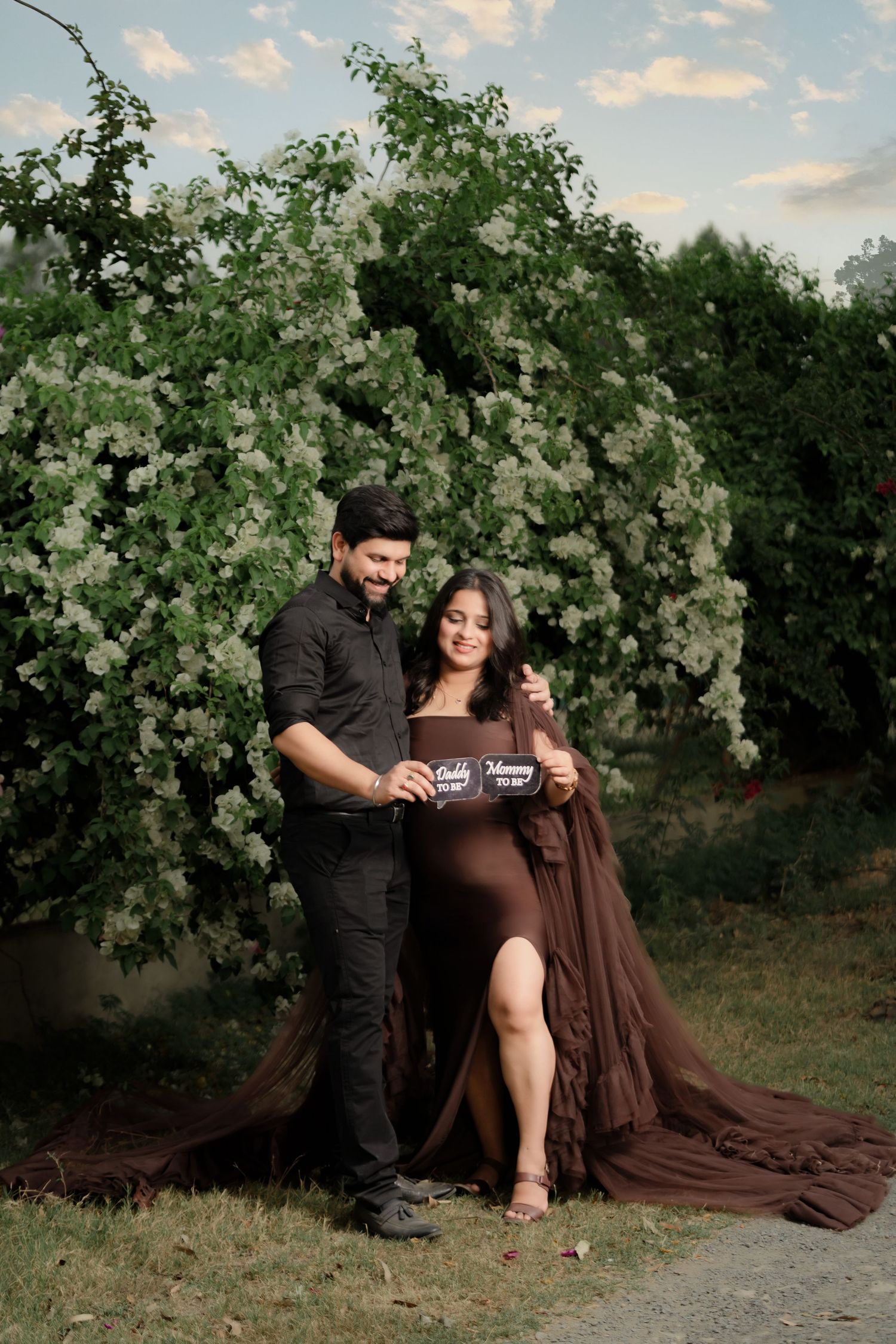 A romantic maternity photo shoot under a blooming tree in a natural outdoor setting with white flowers at sunset.