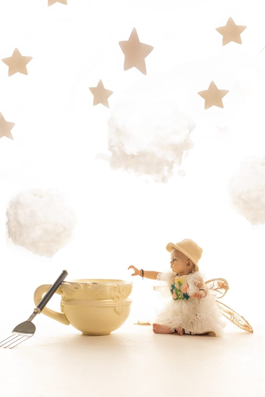 Baby girl in white dress and straw hat sits beside oversized teacup with stars and clouds on white background.