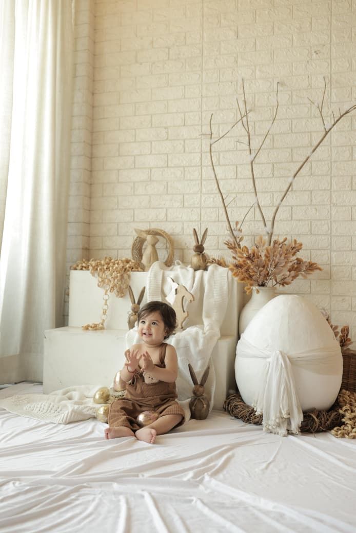 A baby in brown overalls sits on white fabric surrounded by Easter decorations including a large egg and bunny figurines.