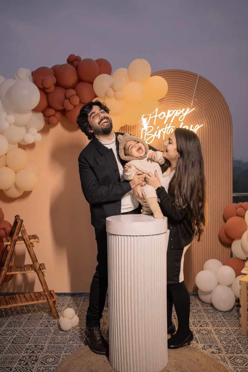 A joyful family celebrates a baby's first birthday with balloon arch decor and a glowing Happy Birthday neon sign.