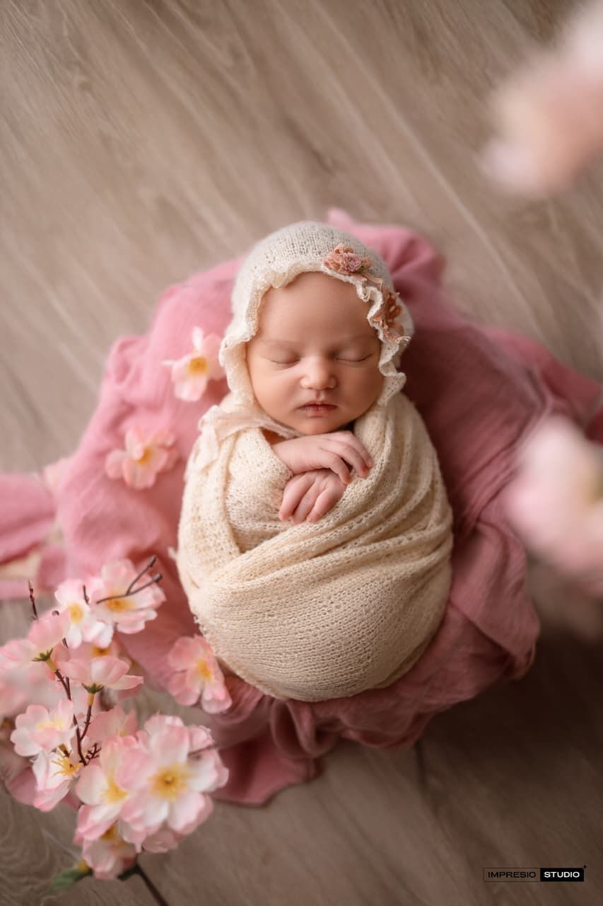Newborn baby swaddled in cream wrap with floral bonnet rests in a bowl surrounded by pink blooms.