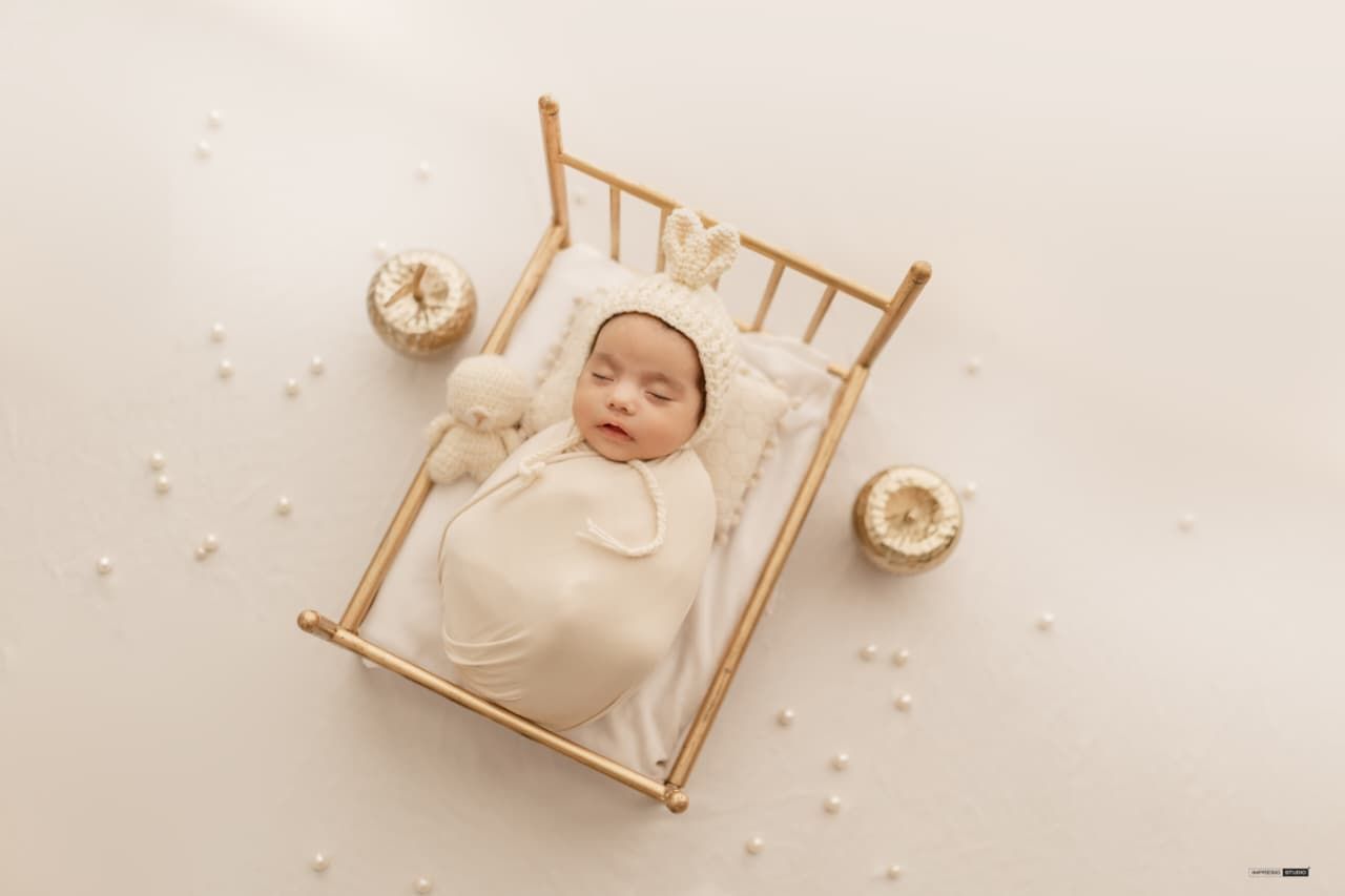 Newborn baby swaddled in cream wrap sleeping in a small wooden crib with soft toys during a studio photoshoot.