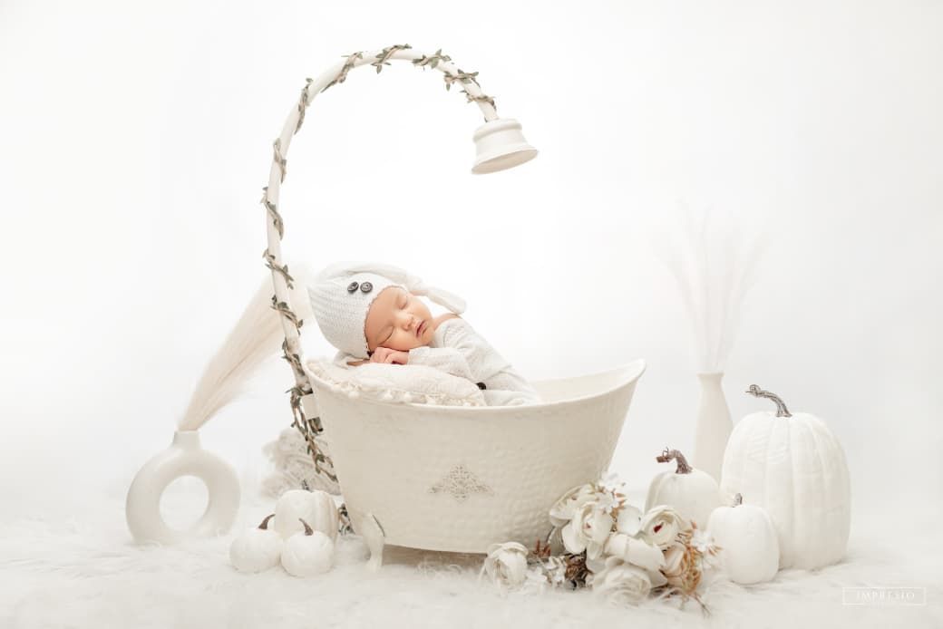 Peaceful newborn baby in white outfit and hat sleeping in decorative white bassinet with pumpkins.