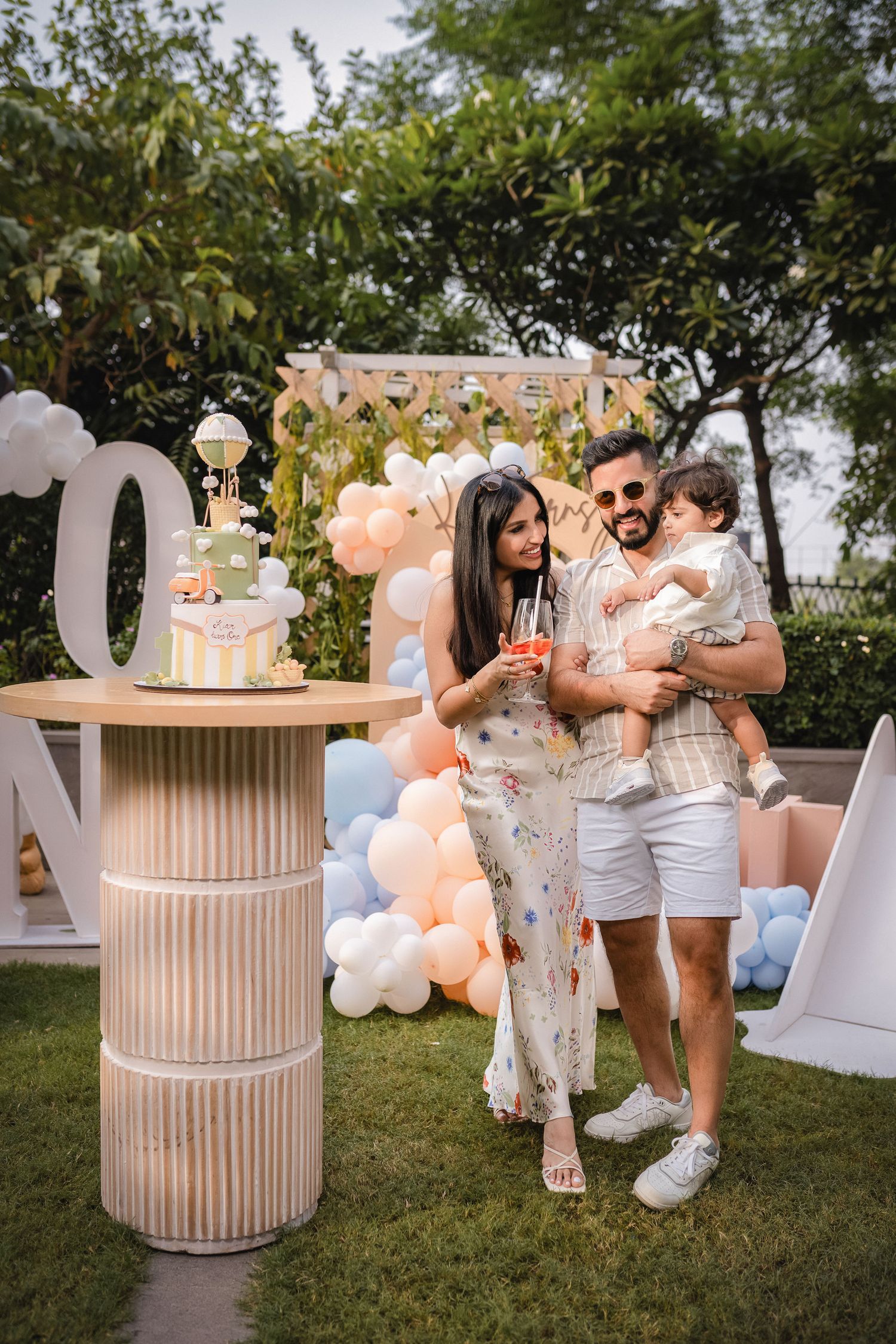 Outdoor party scene with white and peach balloon decorations, rustic barrel tables, and guests celebrating in white attire.