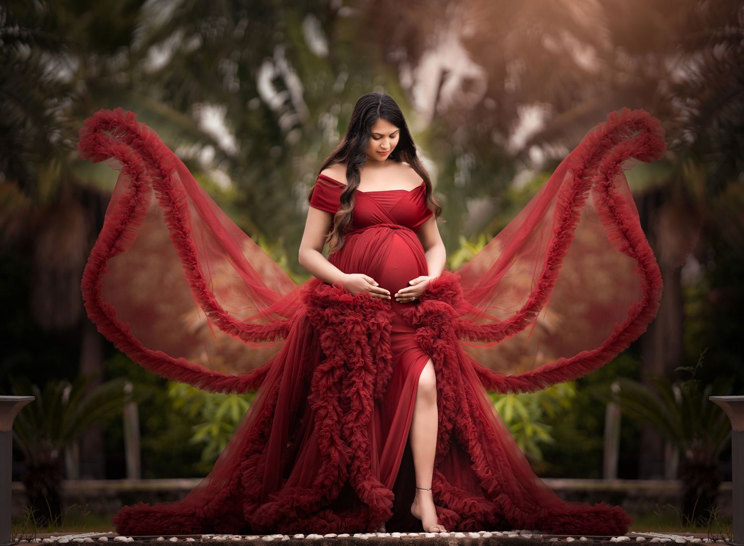 A dramatic maternity photo with a flowing red dress creating wing-like shapes in a garden setting.