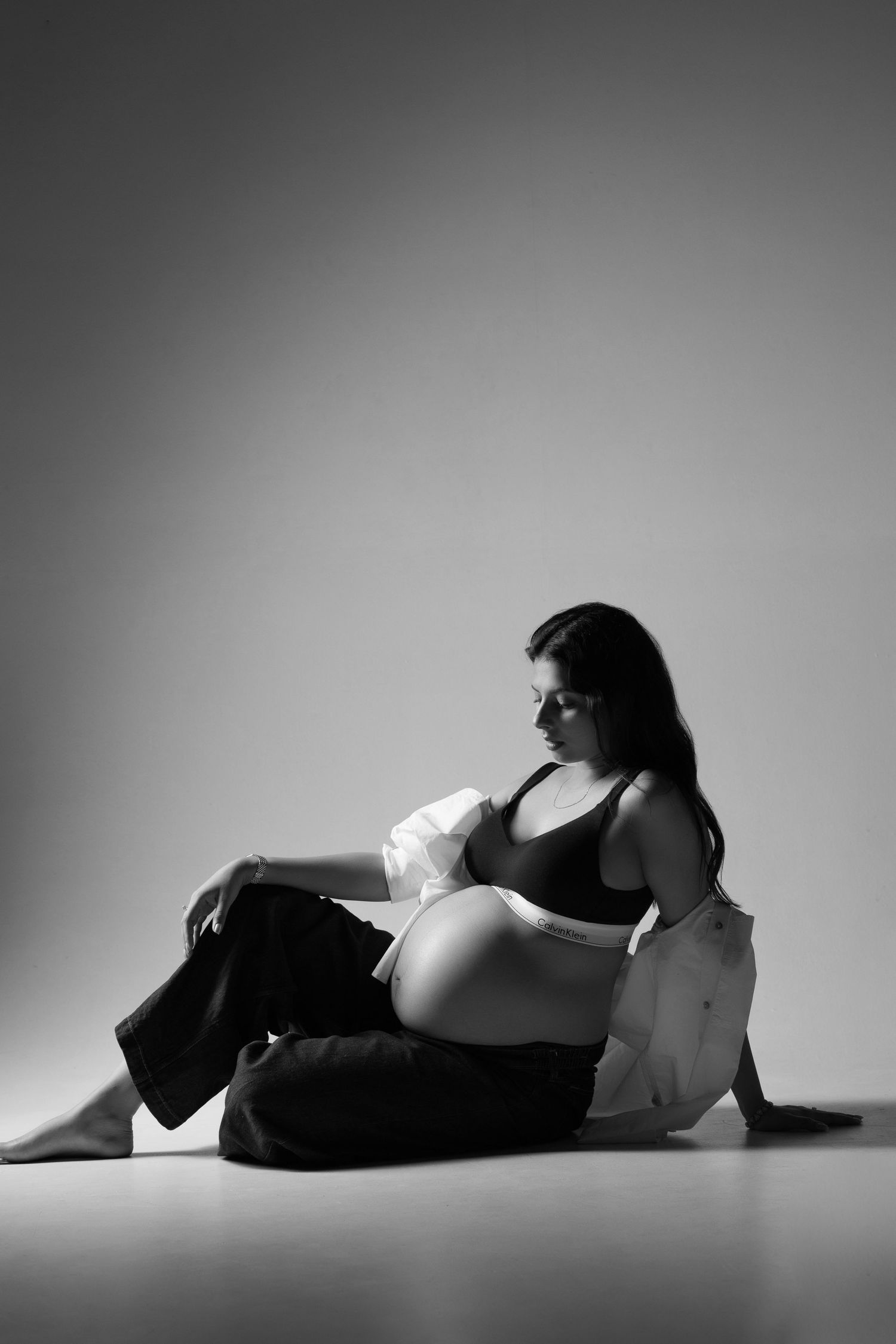 Black and white photo of a pregnant woman sitting on floor, wearing crop top and pants, cradling her baby bump.