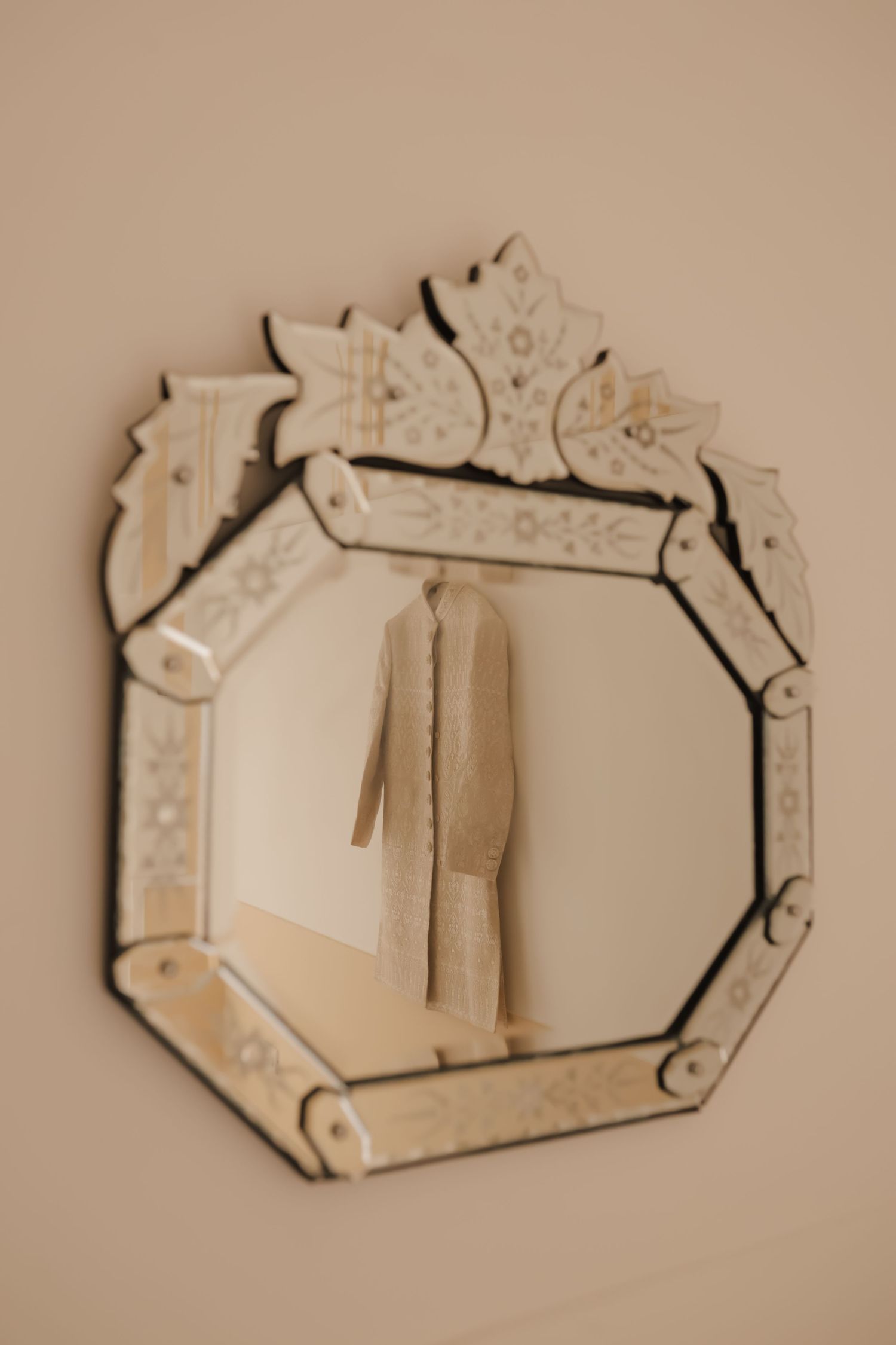 A beige coat hangs on a wall reflected in an ornate vintage Venetian-style mirror with etched glass details.