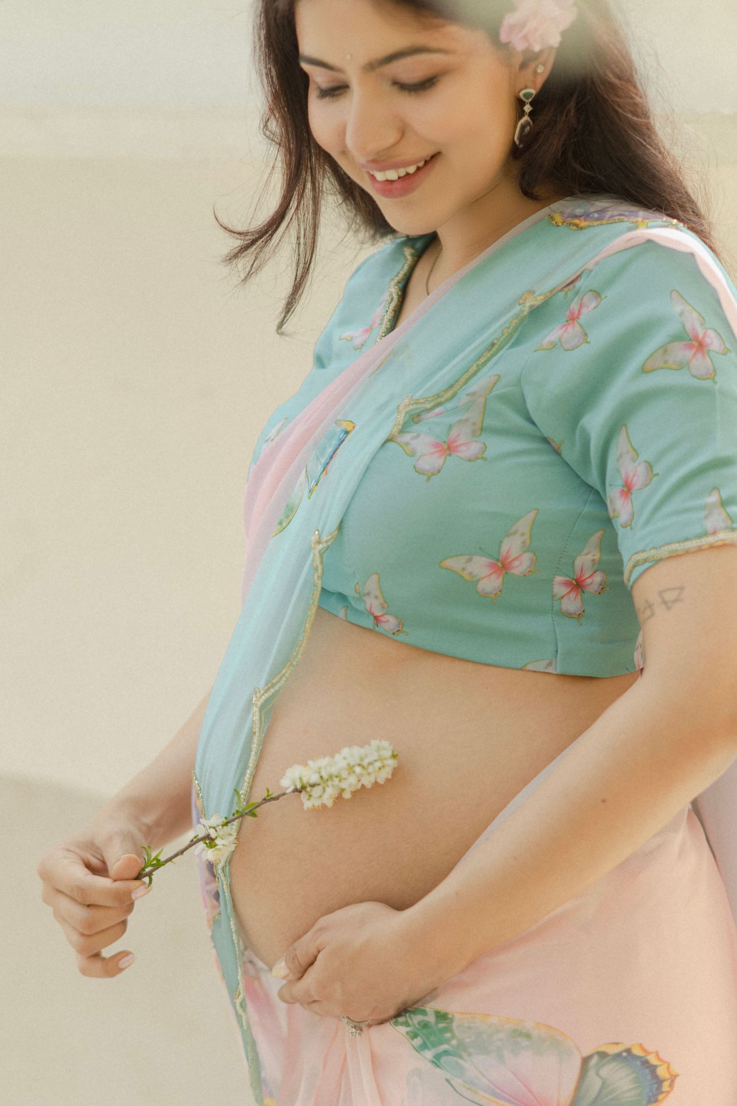 Smiling pregnant woman in teal butterfly crop top and pink saree holding white flowers against her baby bump.