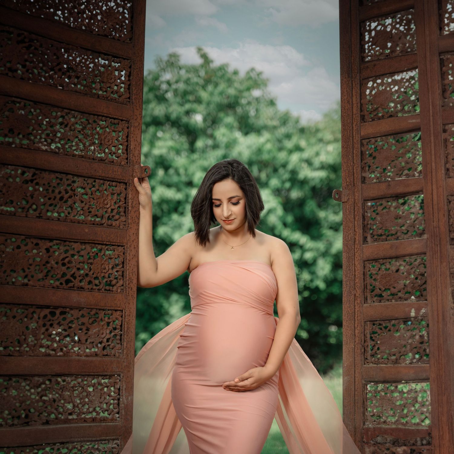 Pregnant woman in pink maternity gown standing in ornate wooden doorway with lush green trees in background.