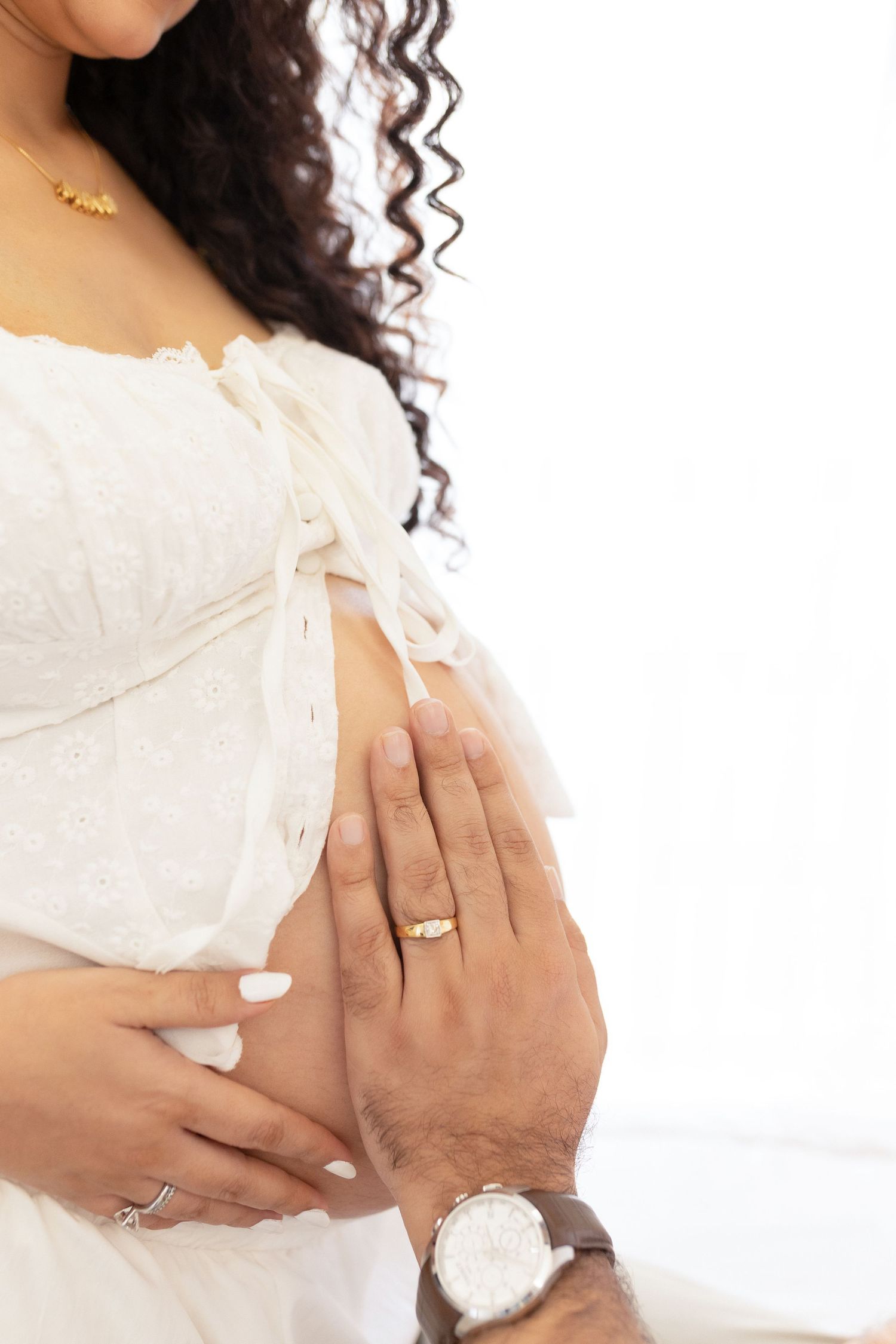 Pregnant woman in white dress holds her belly while wearing a gold ring and showing her curly dark hair.