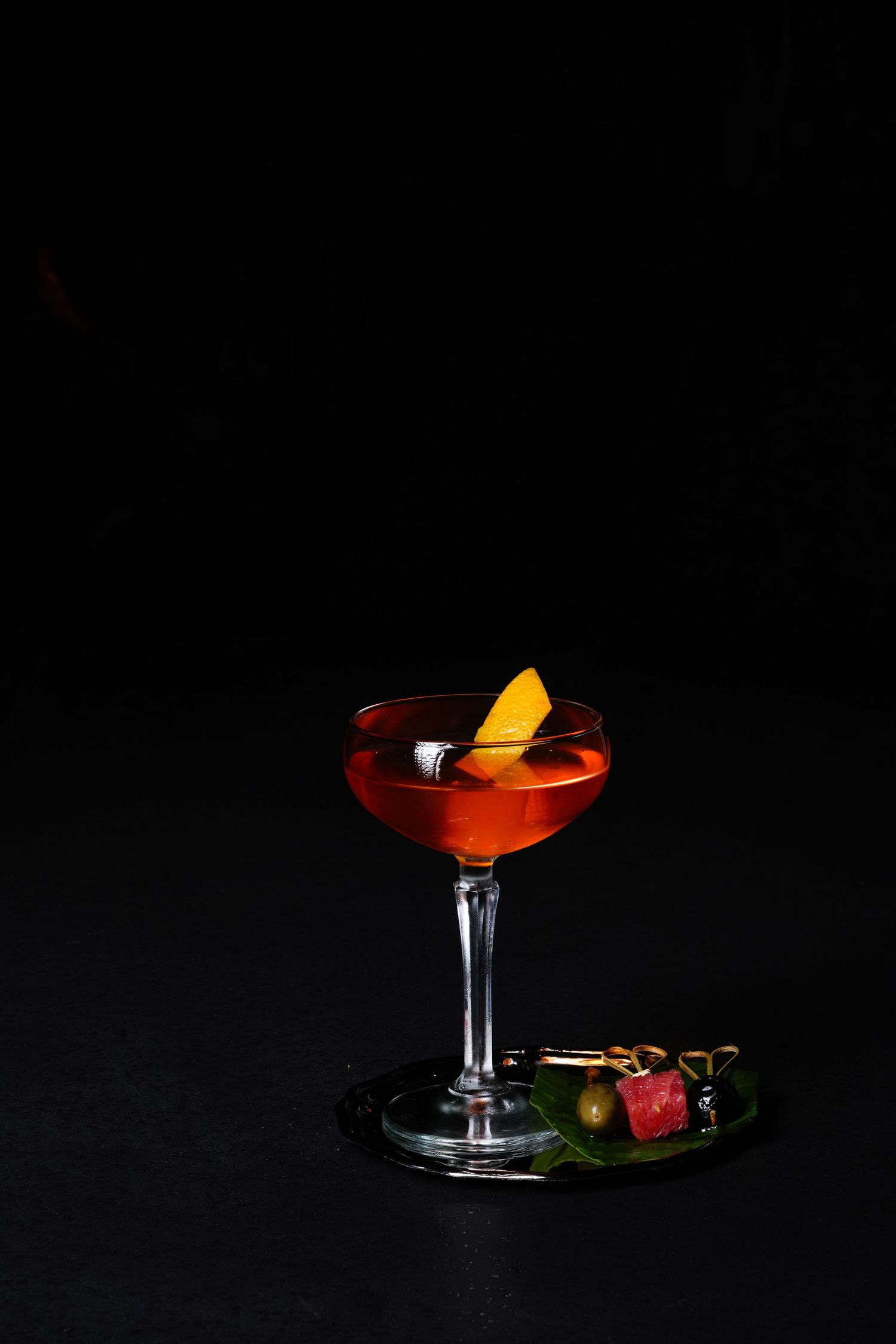 Four elegant cocktail glasses with orange garnishes and dark red drinks photographed against a black background.