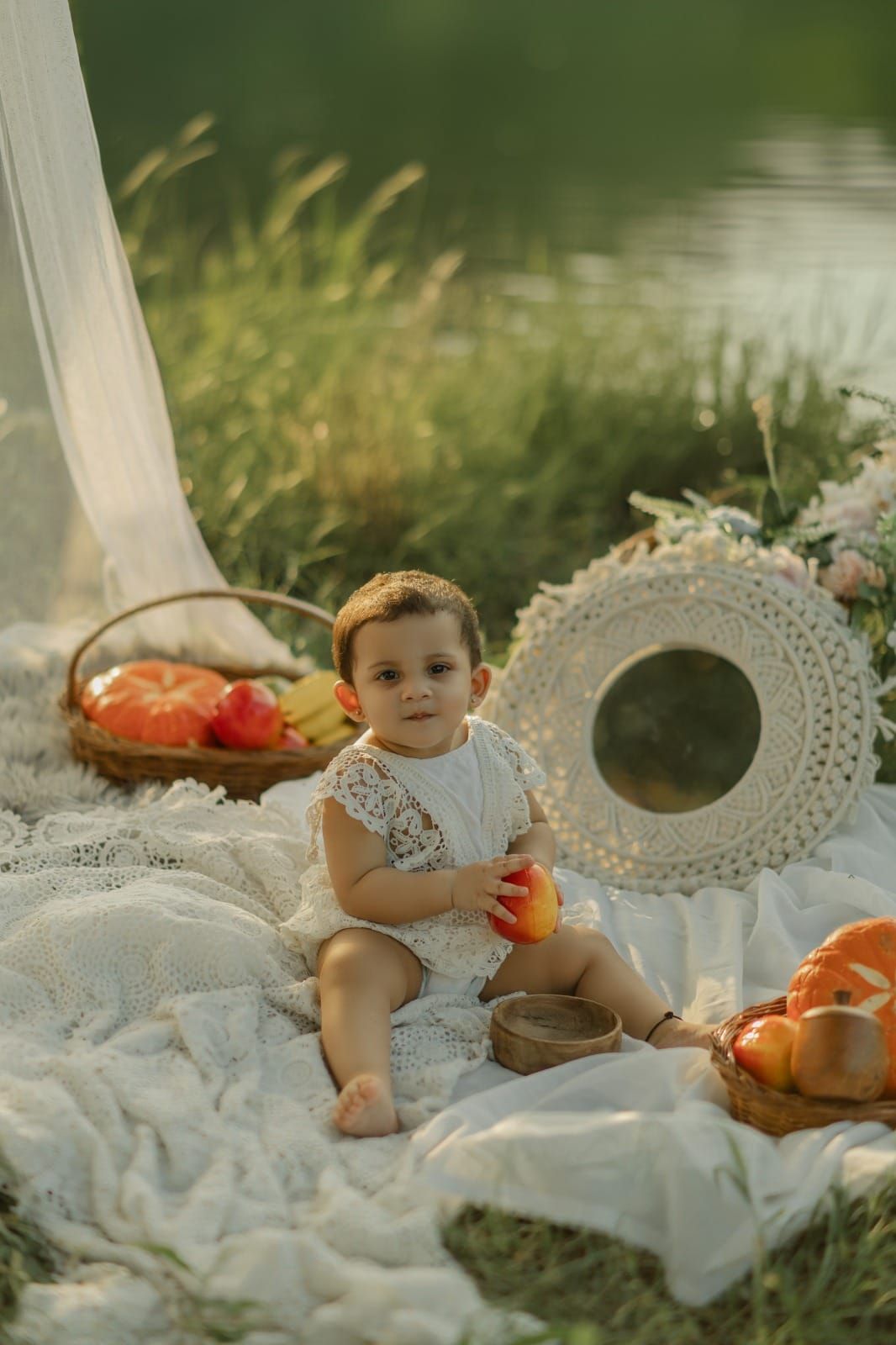 Adorable baby in white lace outfit sitting on blanket outdoors with fruits, flowers, and straw hat near a lake.