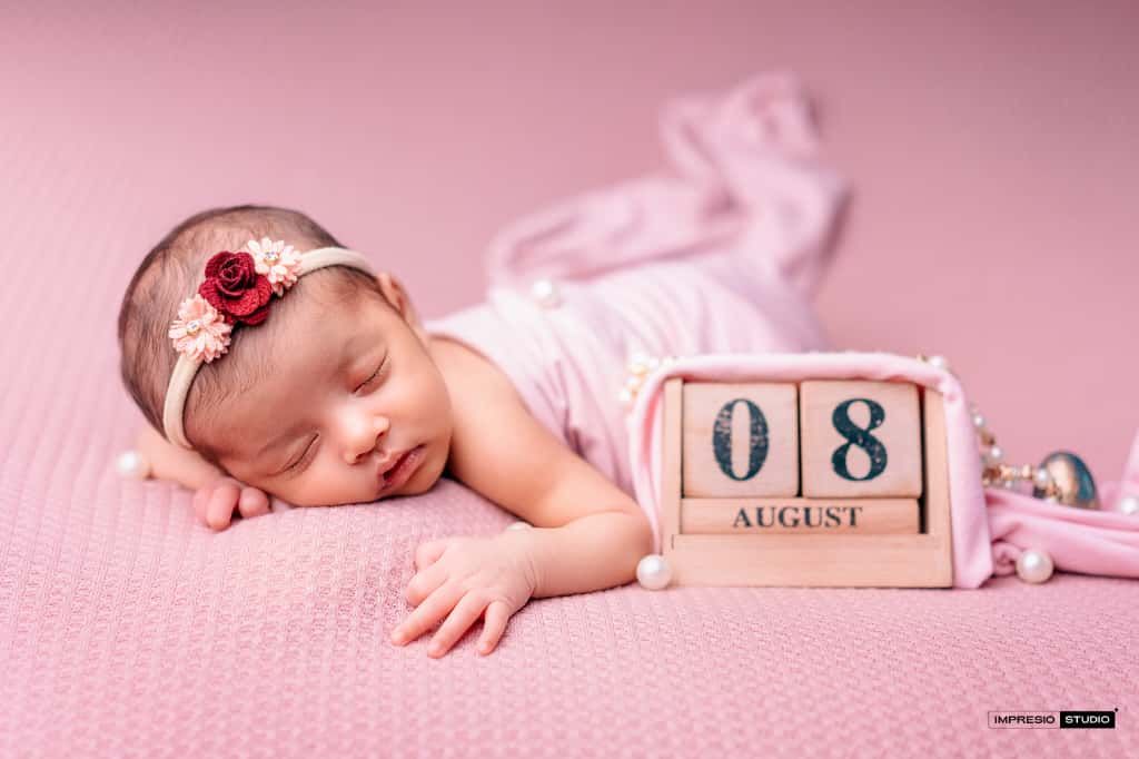 Newborn baby girl sleeping peacefully on pink blanket with floral headband beside August 8th wooden calendar block.