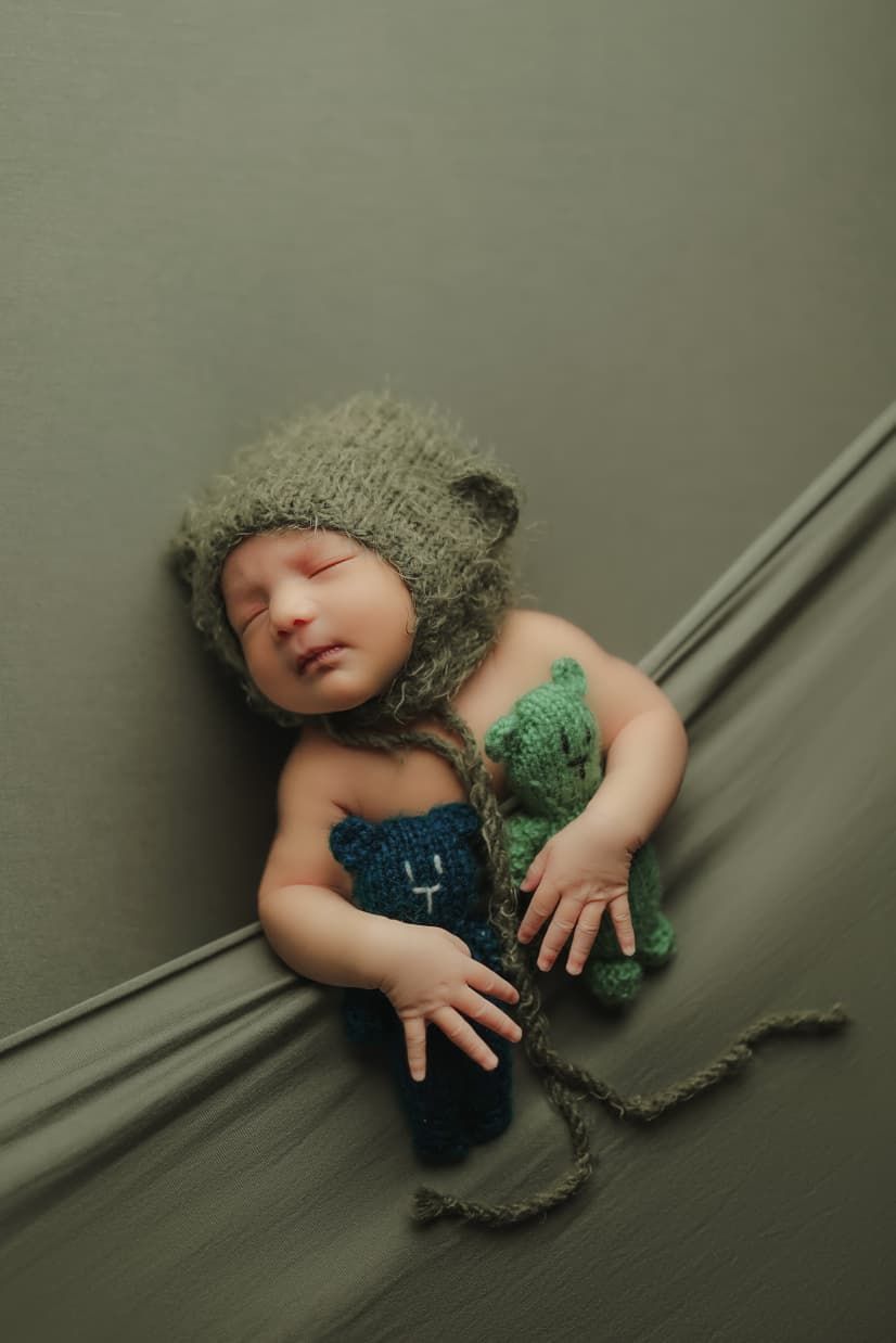 Sleeping newborn baby wearing a knit bear hat holds a small green stuffed animal on a sage green backdrop.