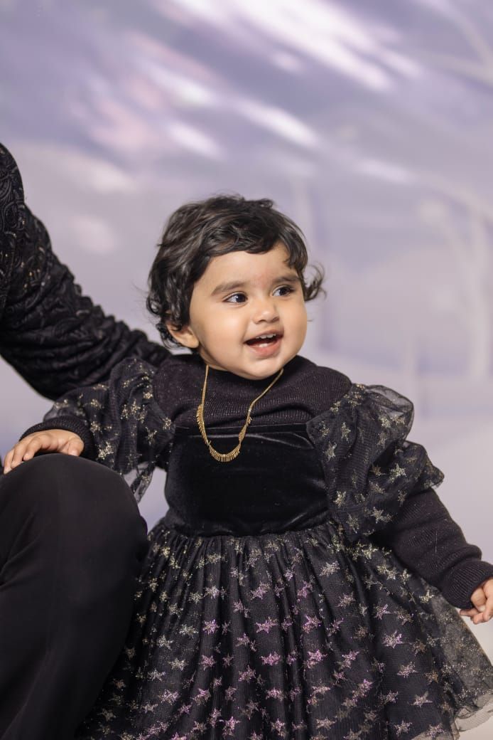 Adorable baby girl in a black star-print dress with gold necklace, smiling joyfully during a studio photo session.