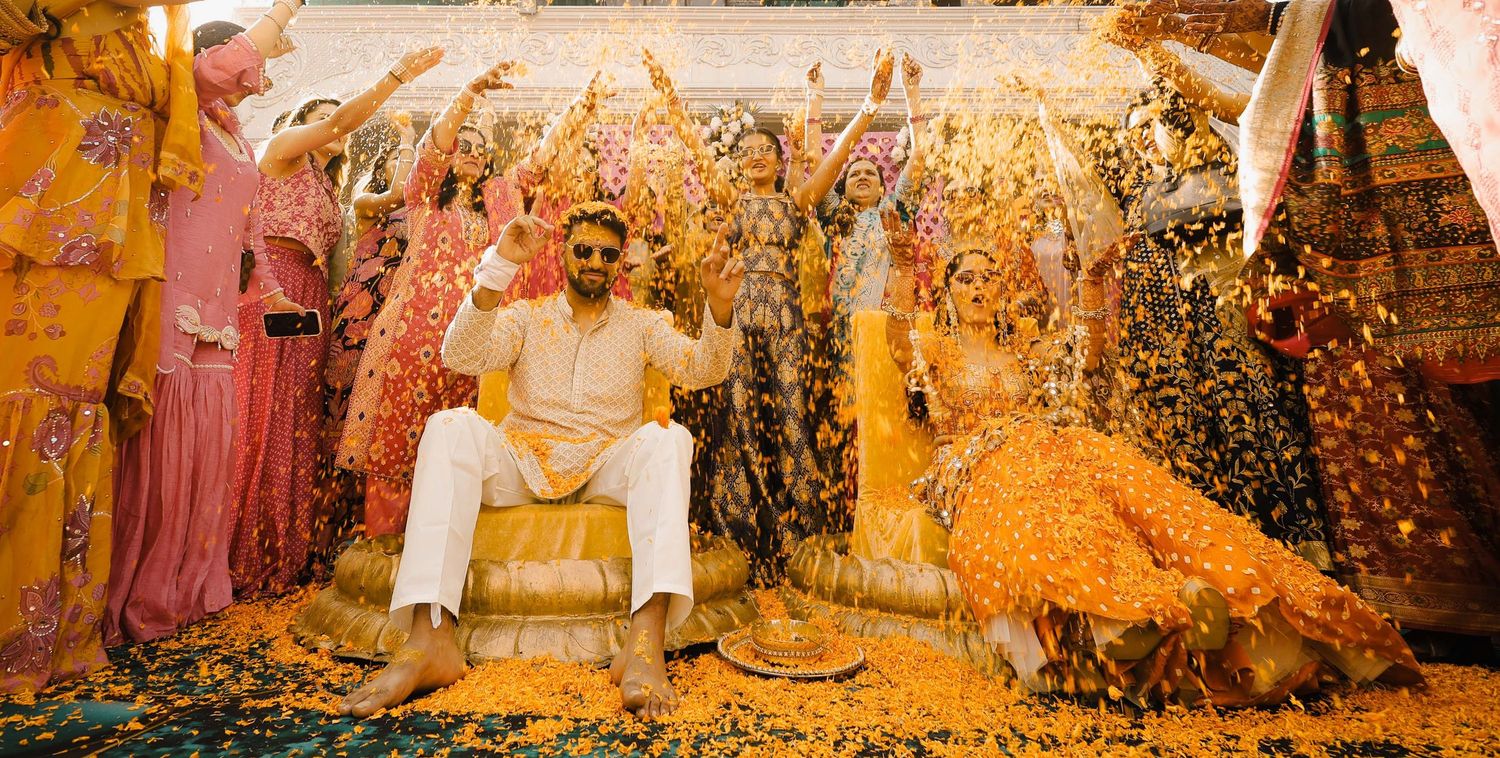 A groom in white sits surrounded by women in colorful attire as flower petals shower down at a vibrant South Asian wedding.