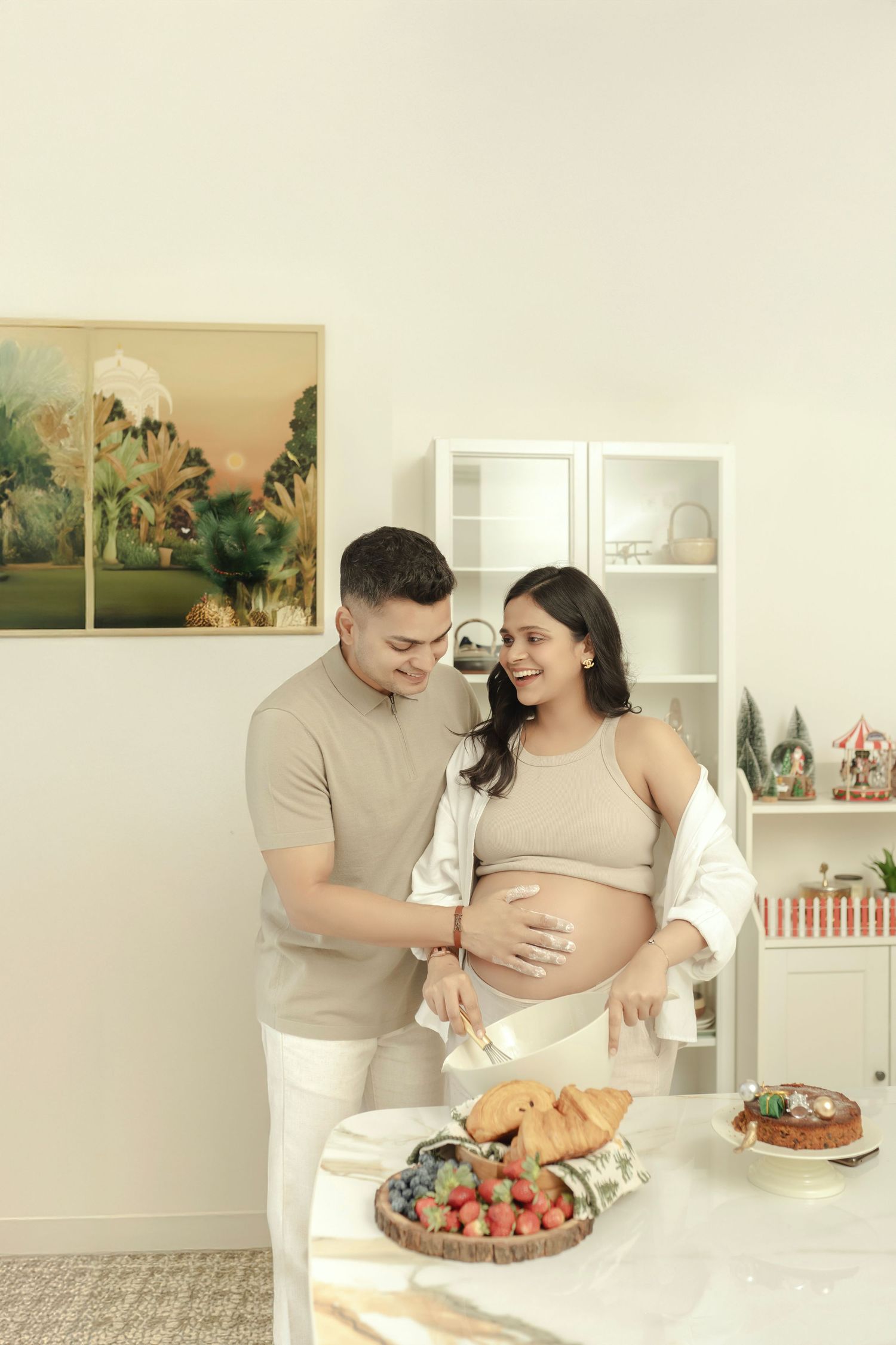 Happy pregnant couple sharing a meal with fresh fruits and croissants in a bright, modern home setting.