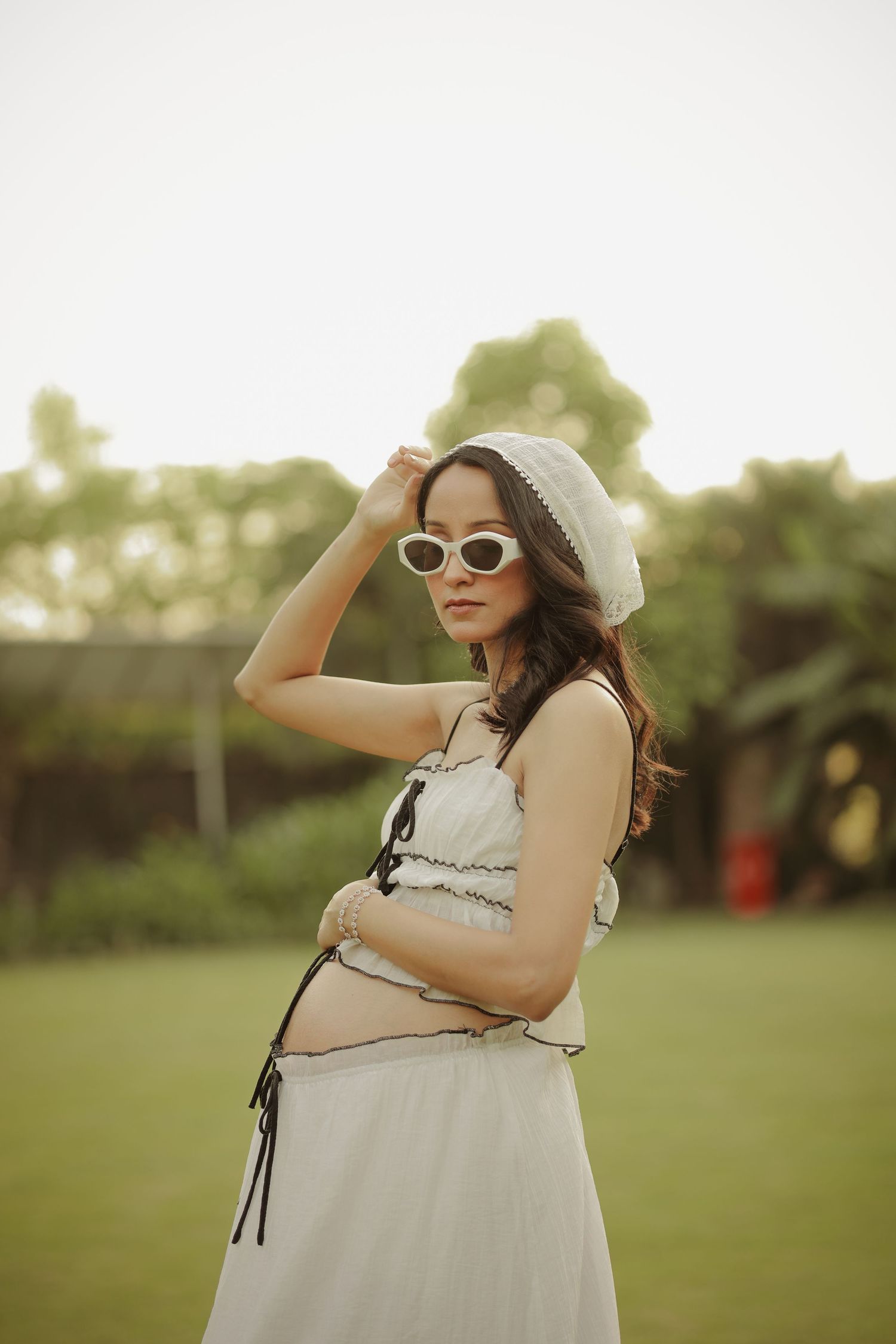 Pregnant woman in white dress, beret, and cat-eye sunglasses posing outdoors on green grass.