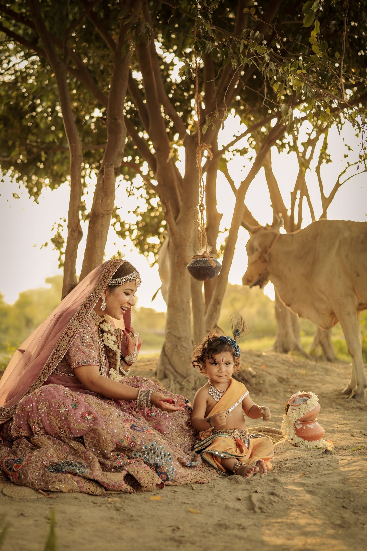 Indian bride in pink lehenga sits with child dressed as Krishna under trees, with a cow in background.
