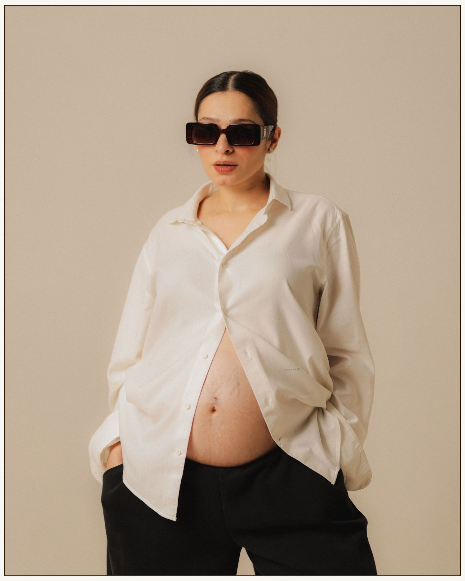 Pregnant woman in white oversized shirt and black pants wearing rectangular sunglasses poses against beige background.