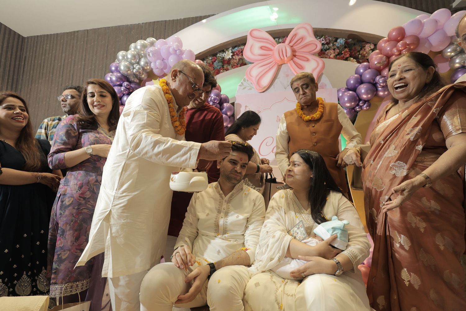 Family celebrates a newborn baby ceremony as an elder blesses a seated couple holding the infant under a pink bow and balloon garland.