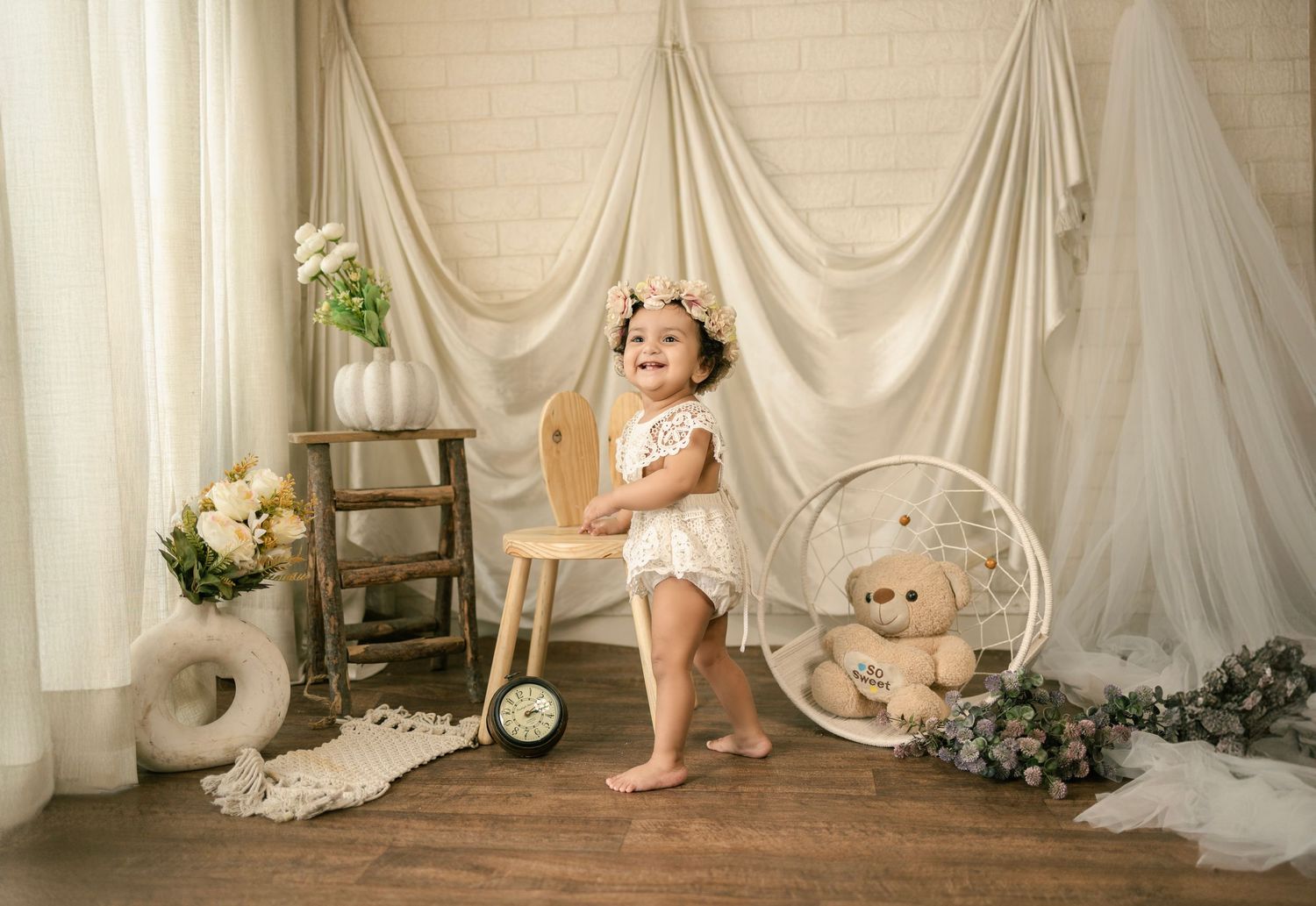 Smiling toddler girl in white lace outfit standing by wooden chair with teddy bear and floral decor backdrop.