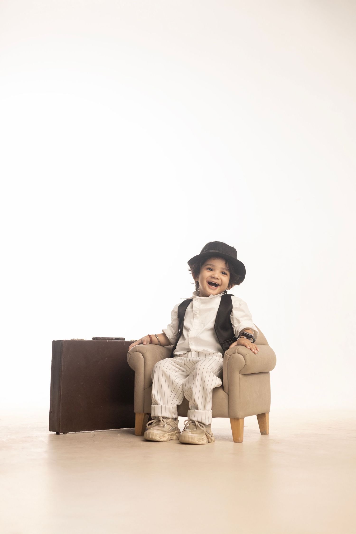 Adorable toddler in white outfit and black hat sits in mini armchair beside vintage brown suitcase, smiling joyfully.