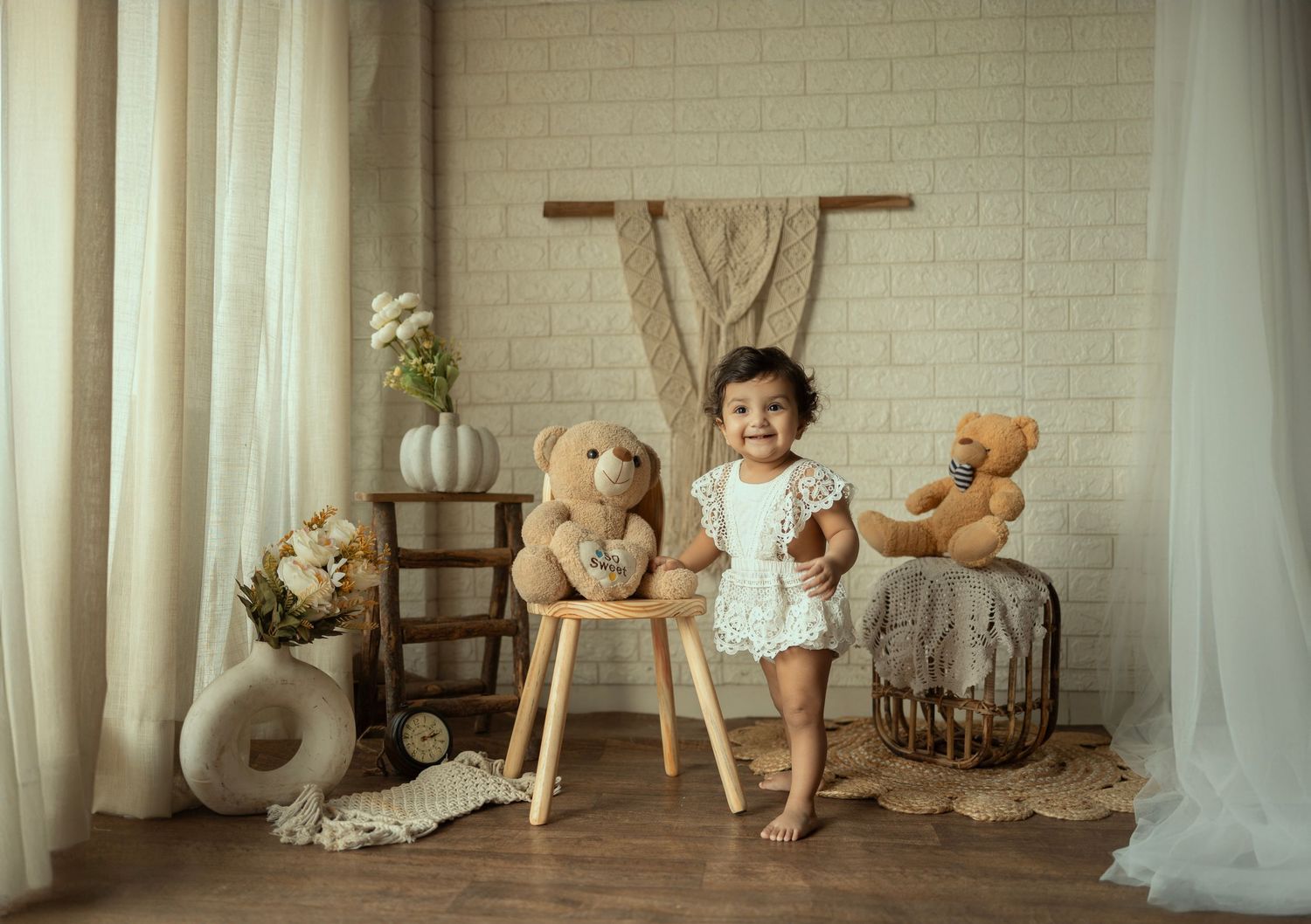 Smiling toddler in white outfit standing between two teddy bears in a cozy, neutral-toned studio photo setup.