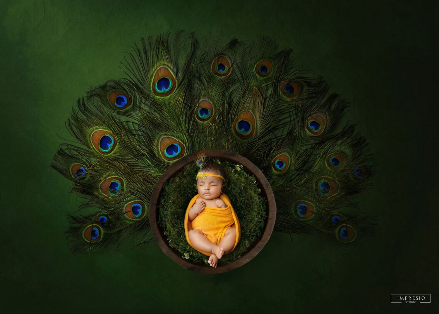 Newborn baby in yellow outfit sleeping in wooden bowl surrounded by colorful peacock feathers on green background.