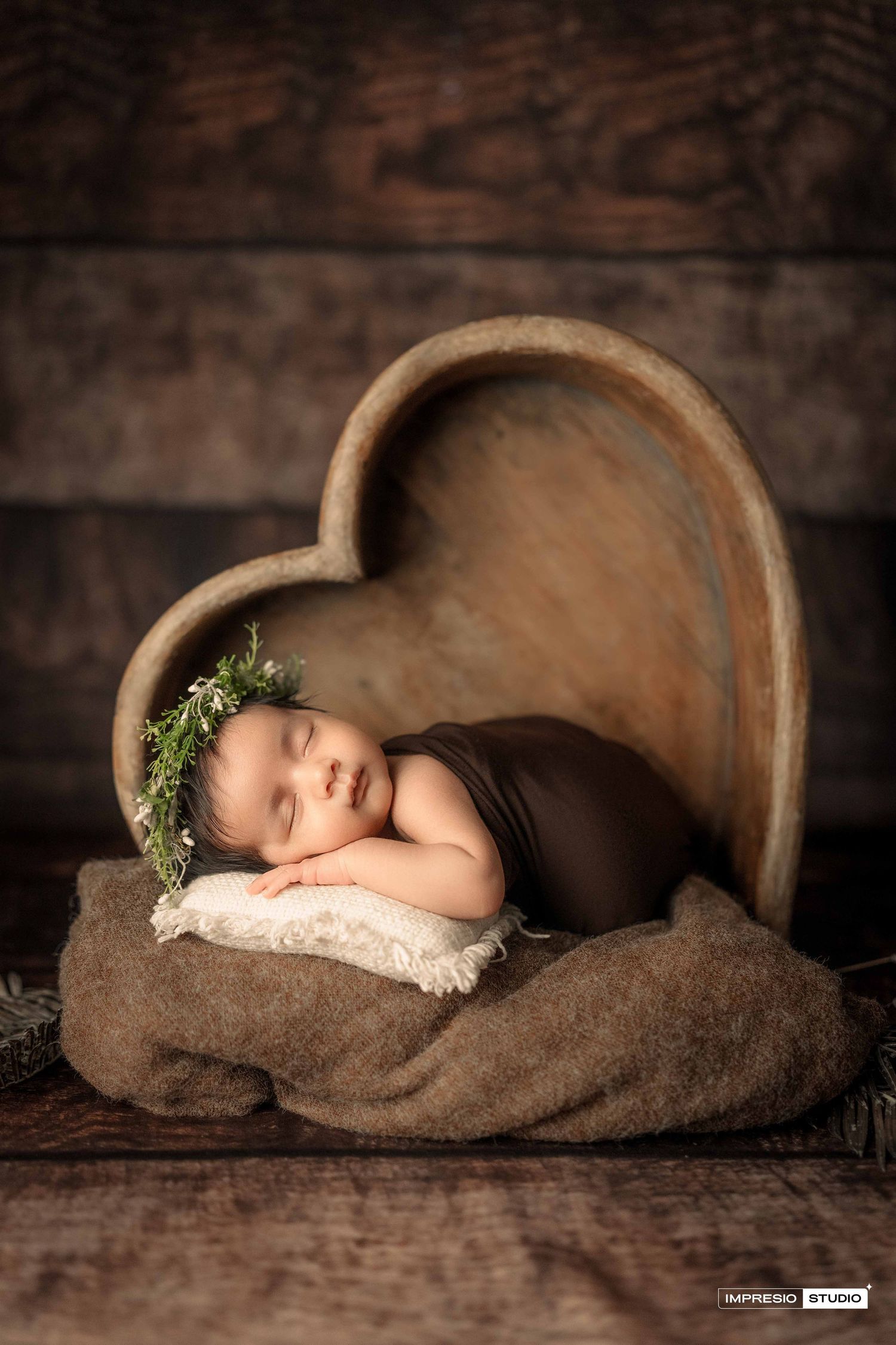Sleeping newborn baby wearing a floral crown nestled in a wooden heart-shaped prop with brown wrap.