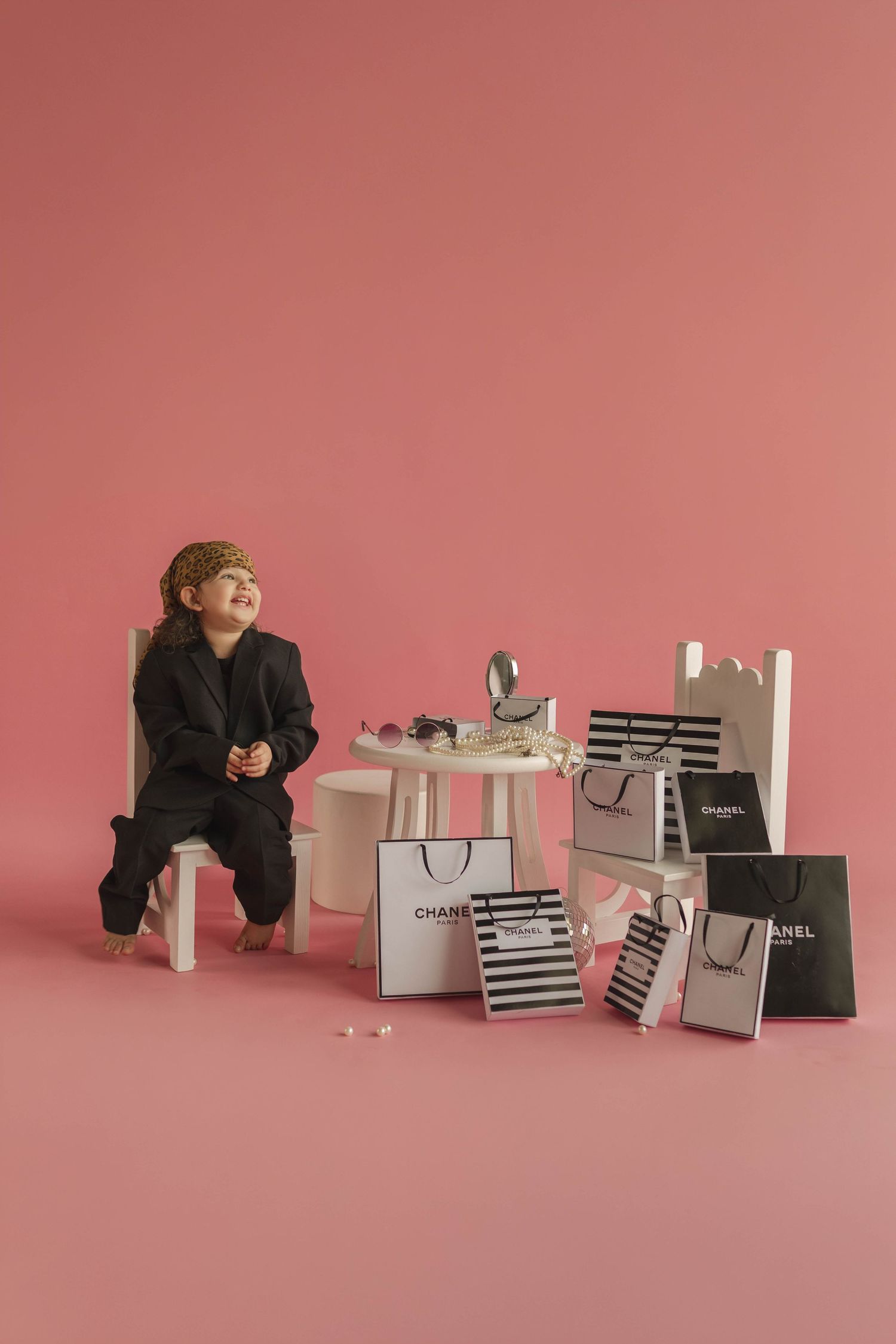 Young child in black suit surrounded by Chanel shopping bags on pink background.