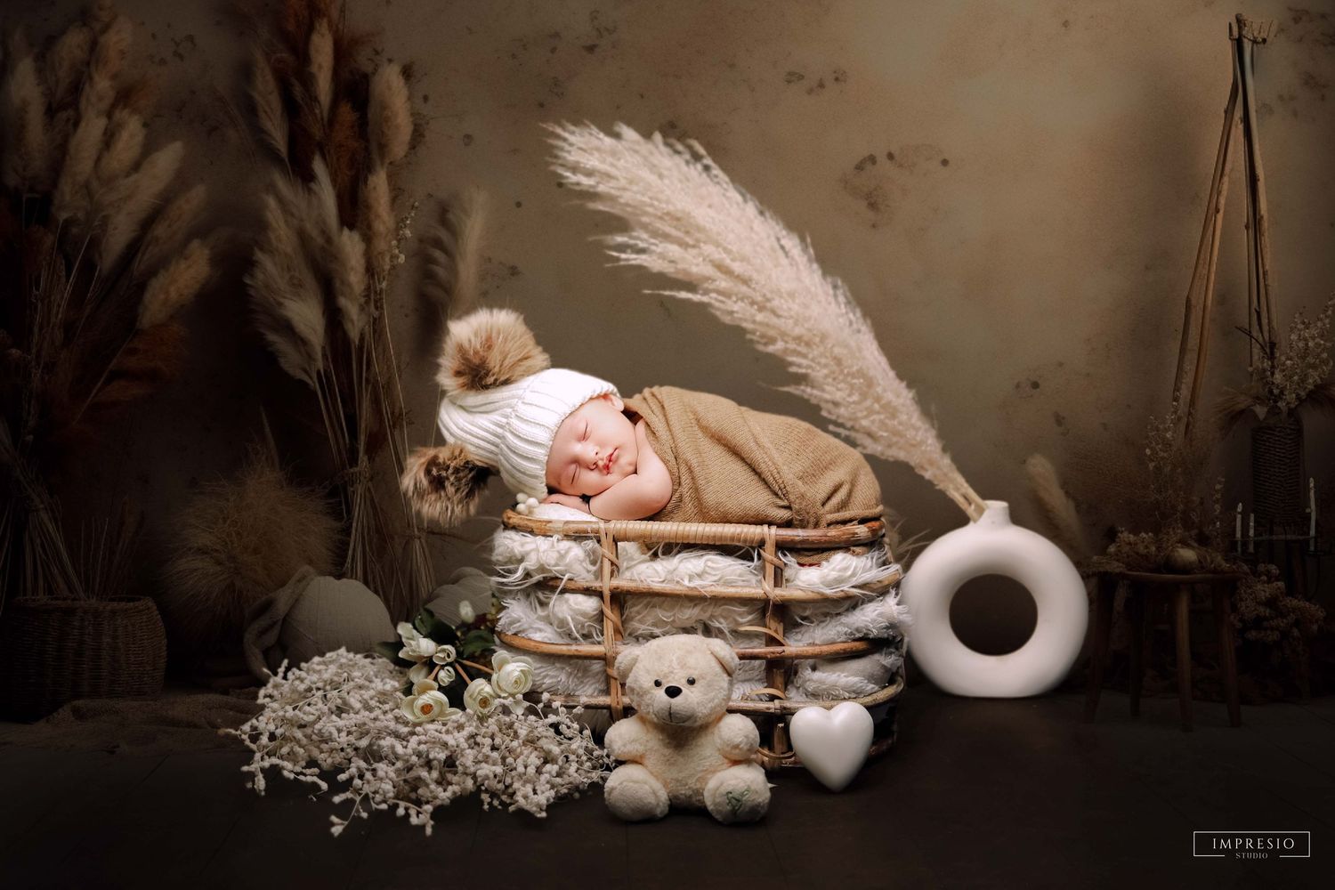 Newborn baby sleeping in rustic wooden crate with teddy bear, pampas grass, and white knit hat in boho studio setting.