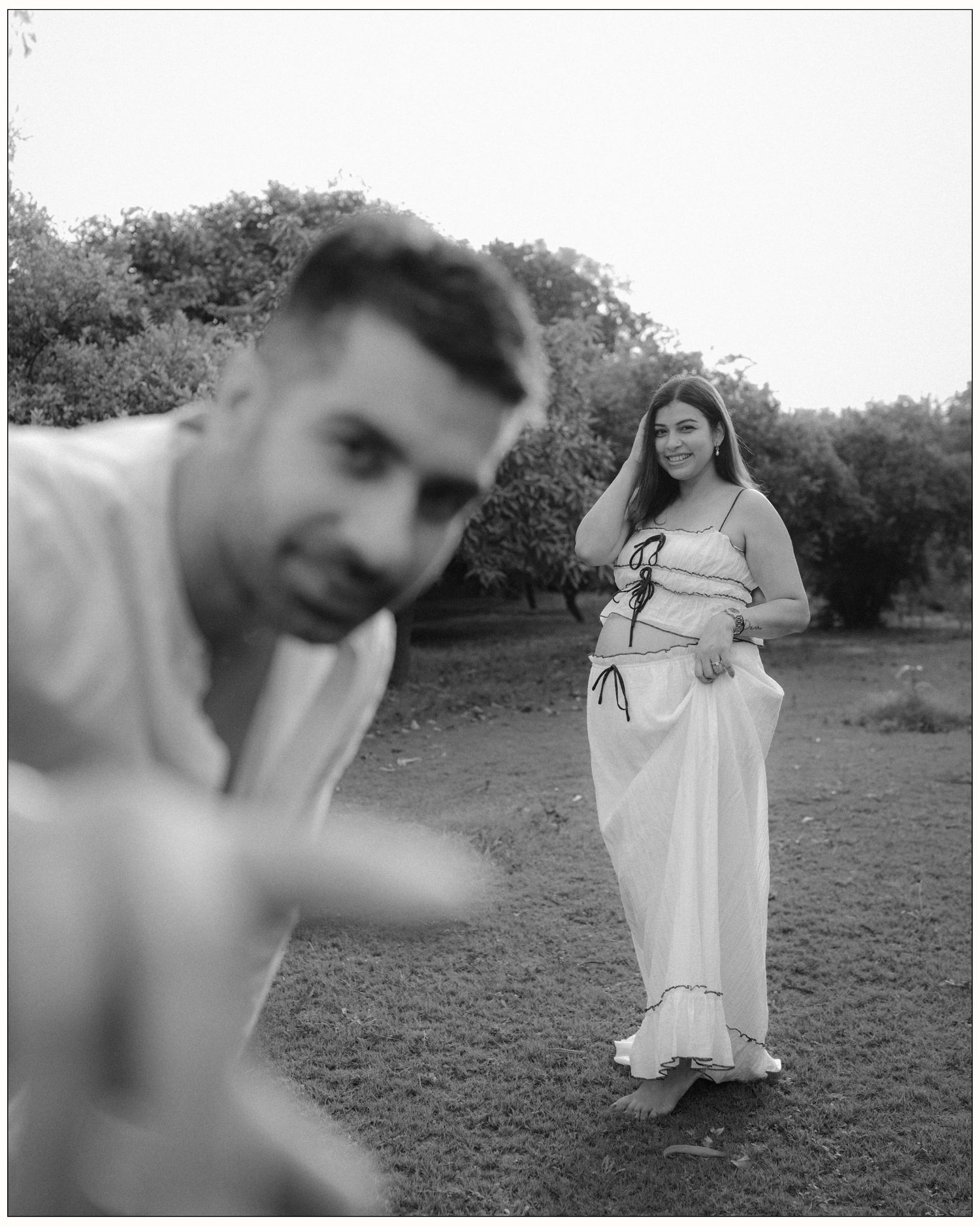 Black and white photo of a couple outdoors, man in foreground, smiling woman in white dress standing among trees.
