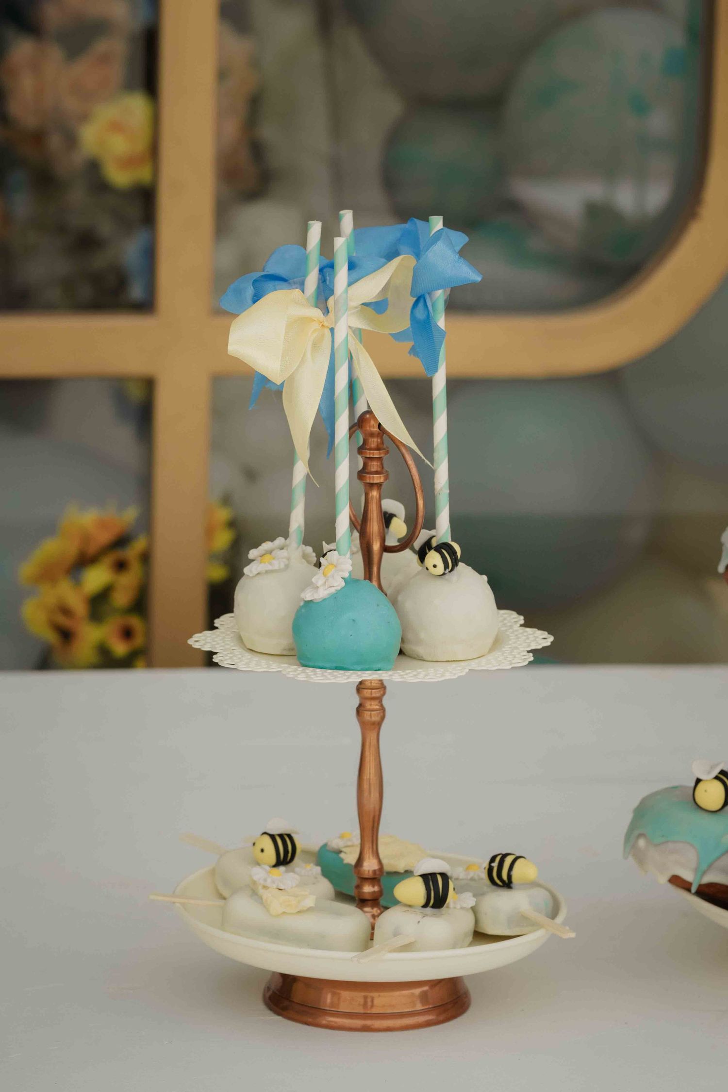 Elegant two-tier cake pop stand with bee-themed treats, blue and white decorations at a baby shower dessert table.
