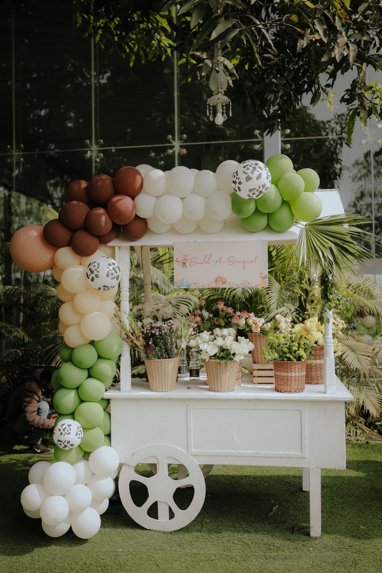 White vintage cart decorated with balloon arch in green, brown, and white, surrounded by potted flowers at an outdoor event.
