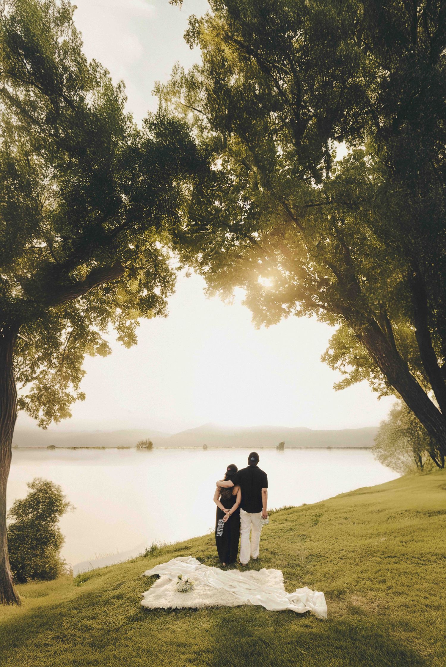 Couple standing under large trees by a serene lake at golden hour, with a white blanket on green grass.