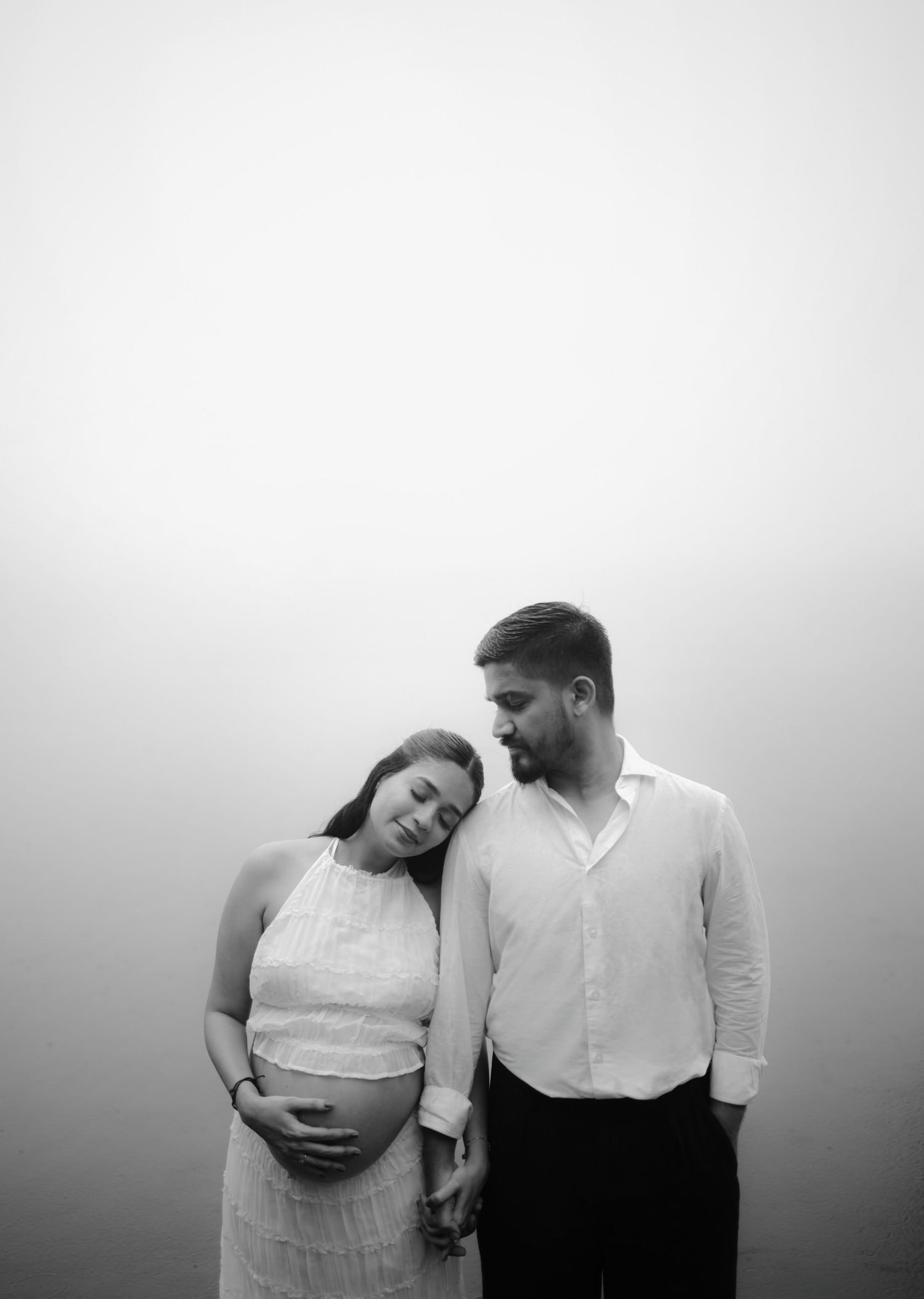 Black and white maternity photo of a couple, woman in white dress cradling baby bump, man in white shirt looking at her tenderly.