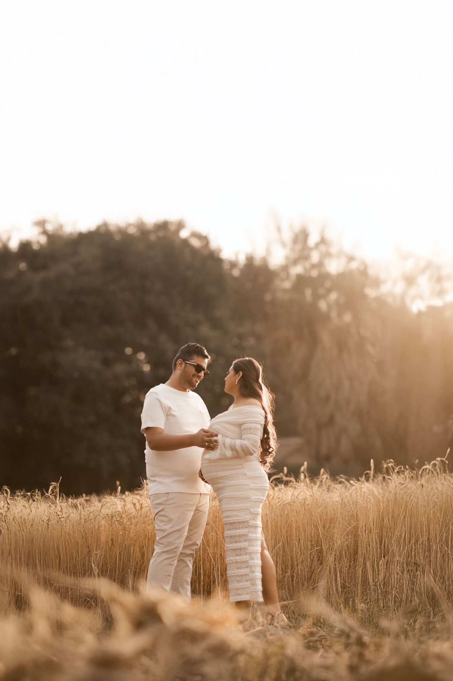 A couple dressed in white embraces in a golden meadow for maternity shoot with husband at sunset.