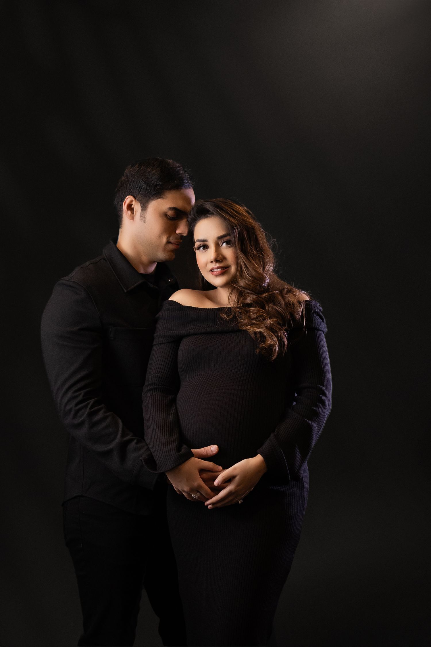 Intimate maternity photo series with couple wearing black against dark background shows tender moments and embraces.