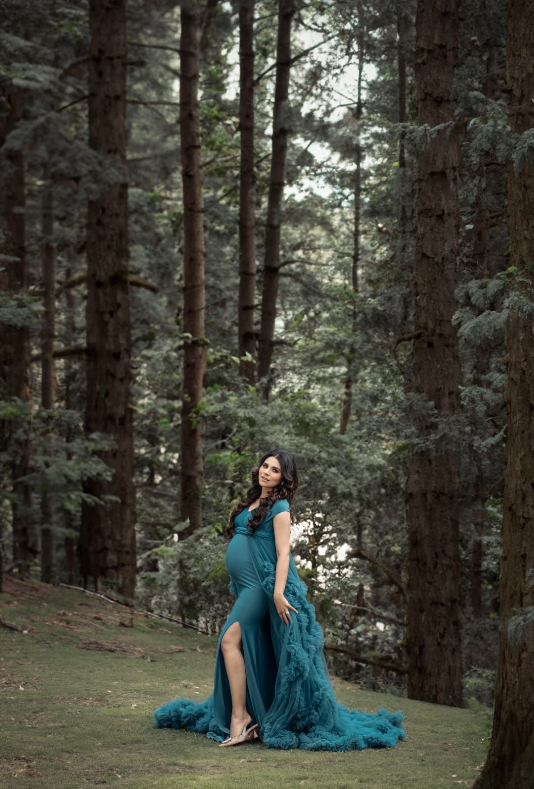 Pregnant woman in teal ruffled gown poses gracefully among tall trees in a lush forest maternity photoshoot.