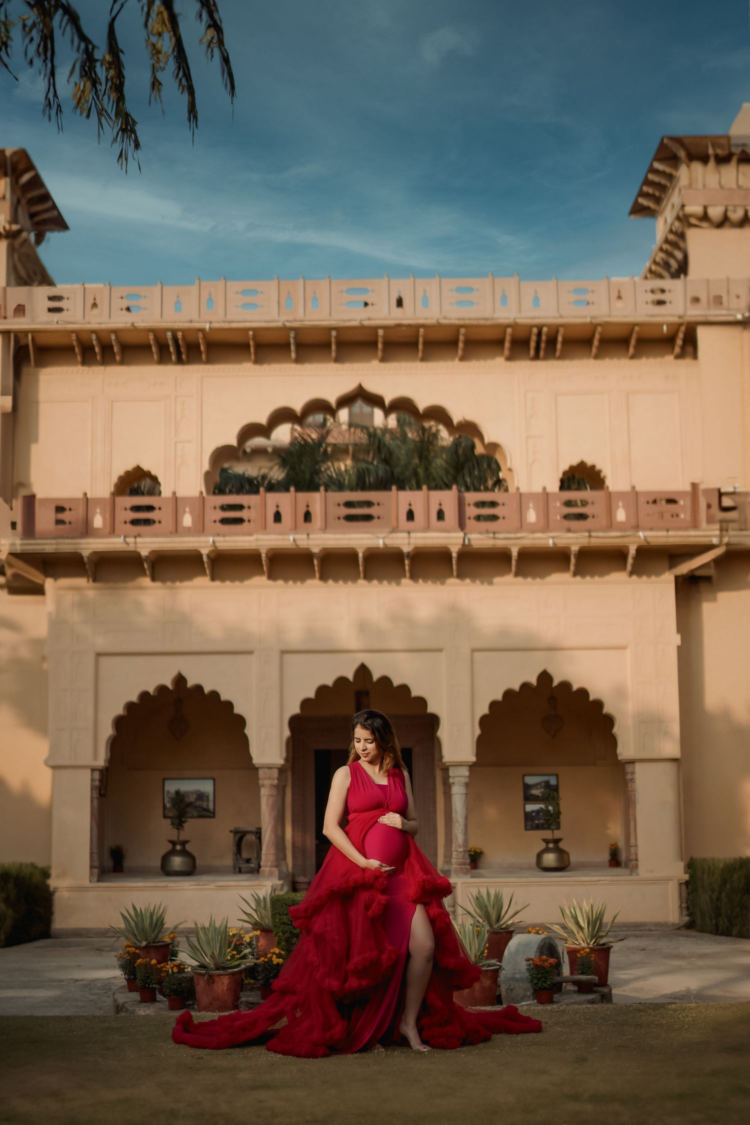 Pregnant woman in red gown poses before ornate Rajasthani palace with arched corridors and blue sky.
