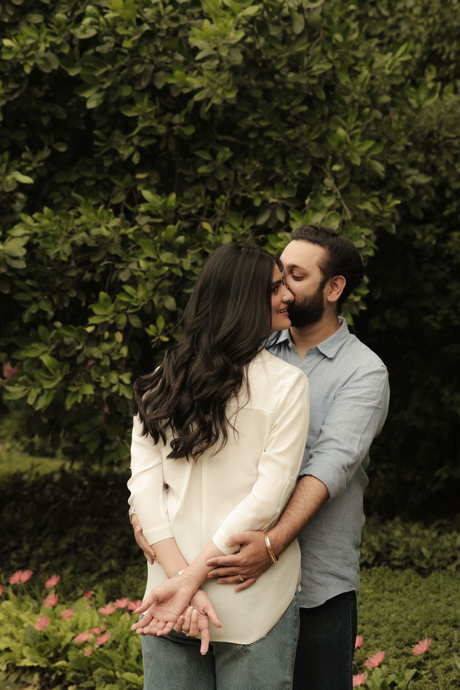 Couple embracing in a lush garden, man kissing woman's cheek, both wearing casual blue and white outfits.