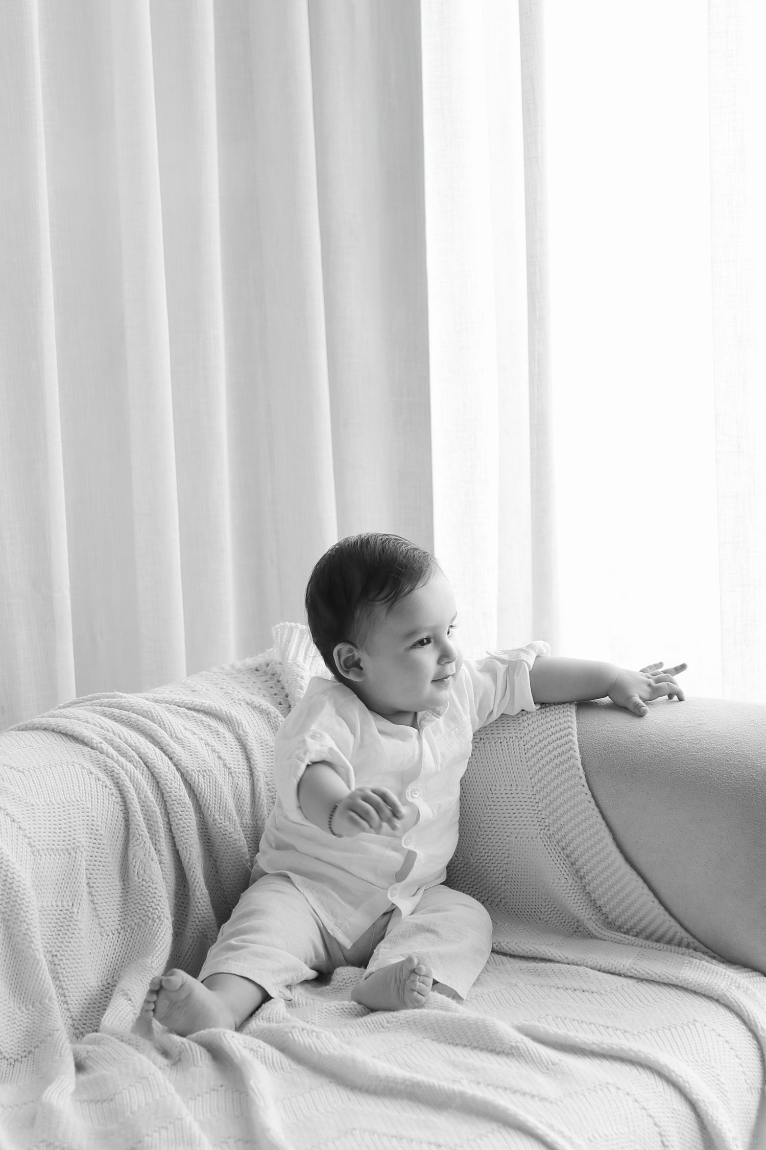 Black and white photo of a curious baby sitting on a sofa, reaching out toward a bright window with curtains.