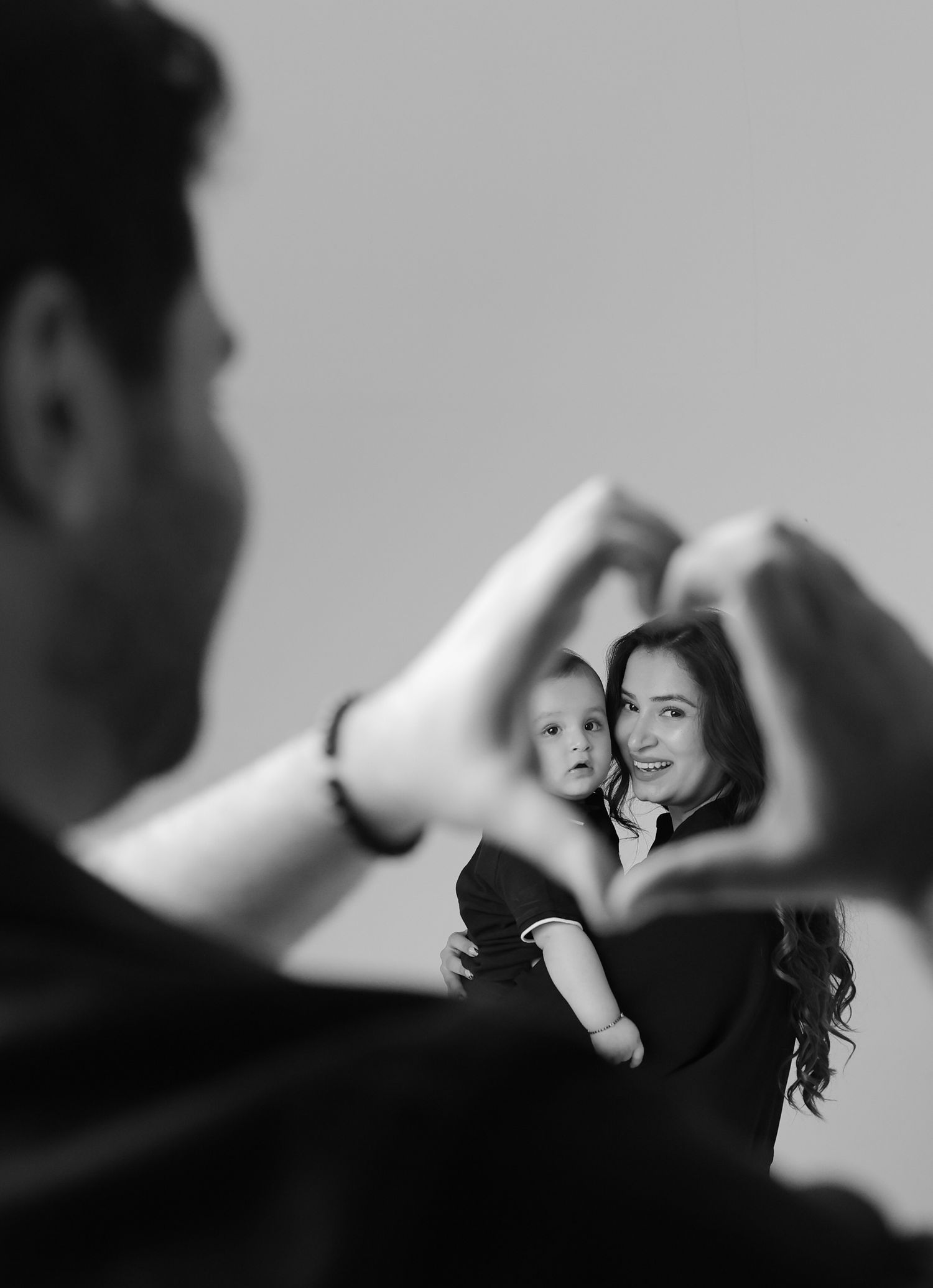 Black and white photo of a man forming a heart shape with his hands, framing a smiling woman holding a baby.