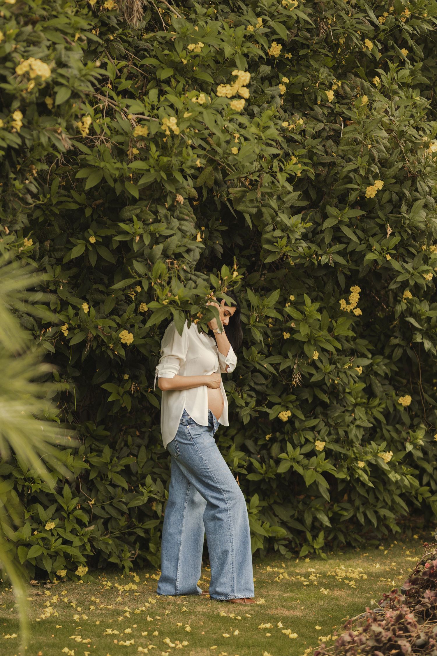 Woman in white shirt and wide-leg jeans standing in lush garden with yellow flowering bushes.