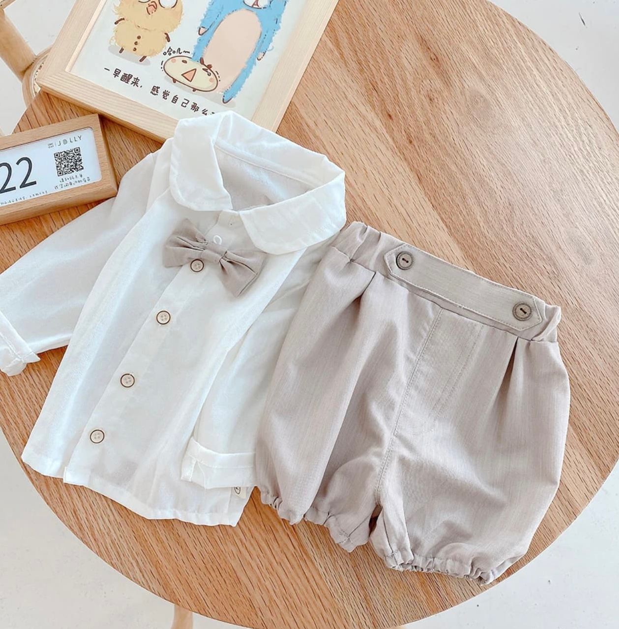 Classic baby outfit set with white collared shirt with bow tie and light beige bloomers shorts on wooden surface.