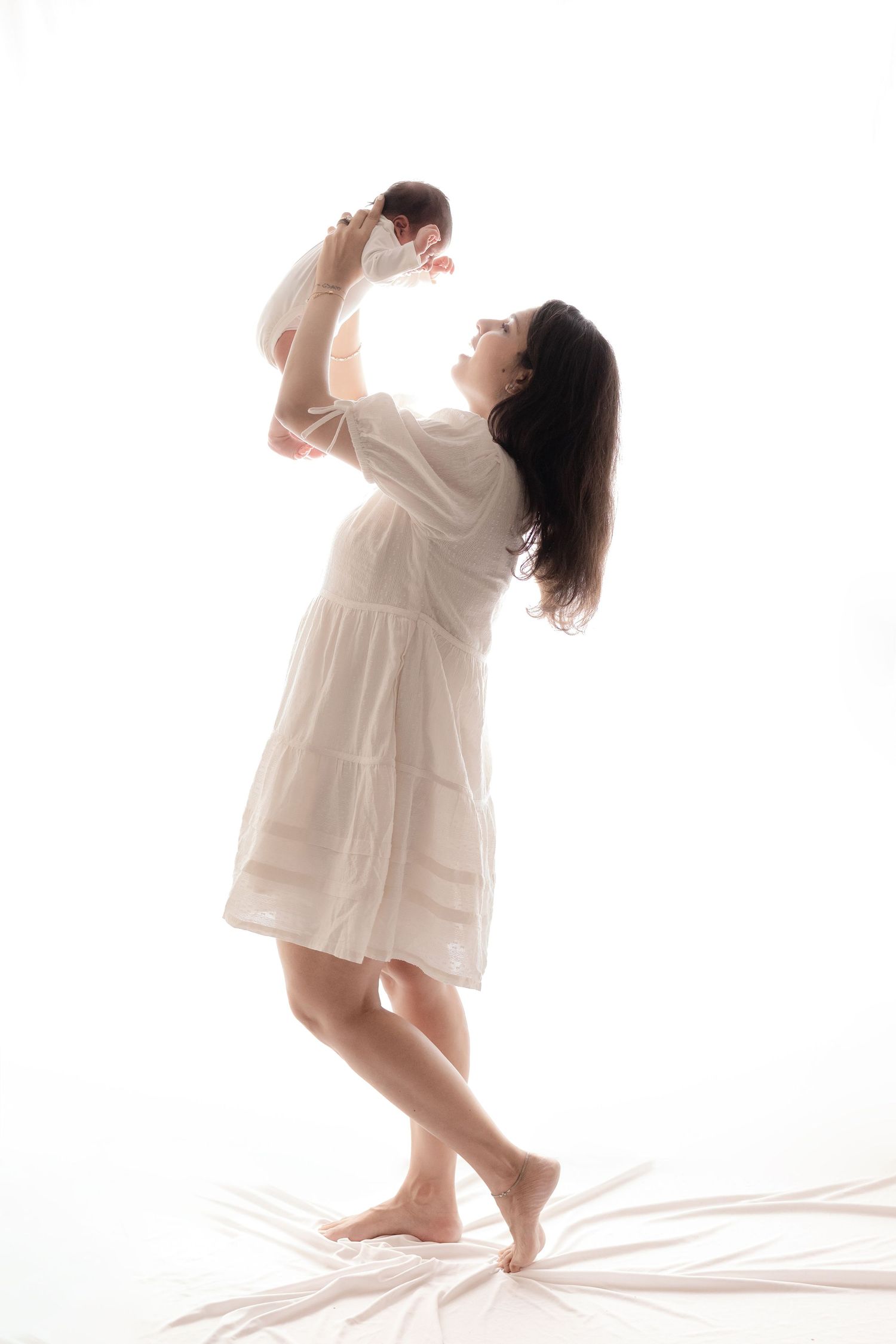 A barefoot mother in a white dress lifts her newborn baby overhead, both illuminated against a bright white background.