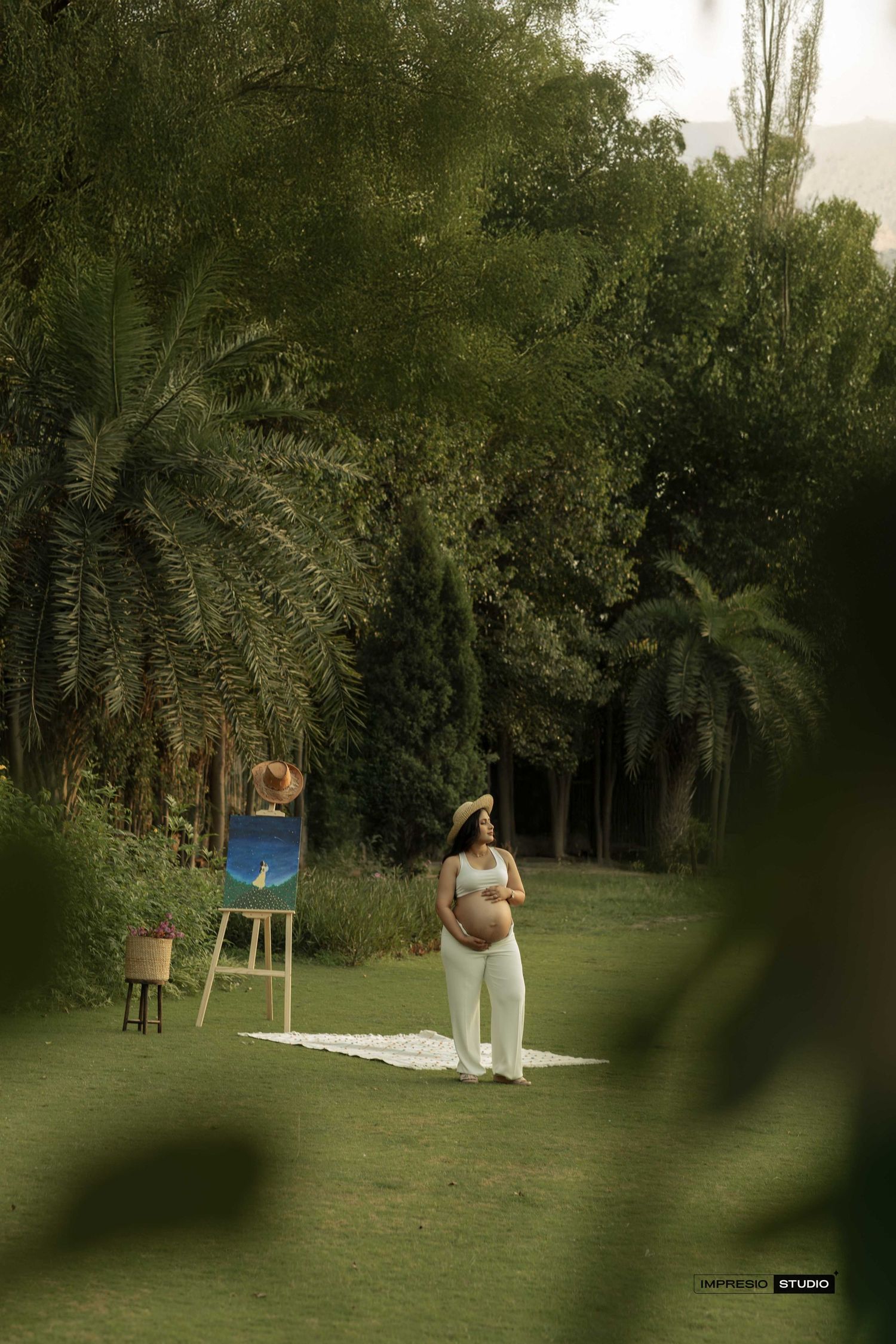 Pregnant woman in white outfit stands on lawn beside an easel painting in a lush green garden setting.