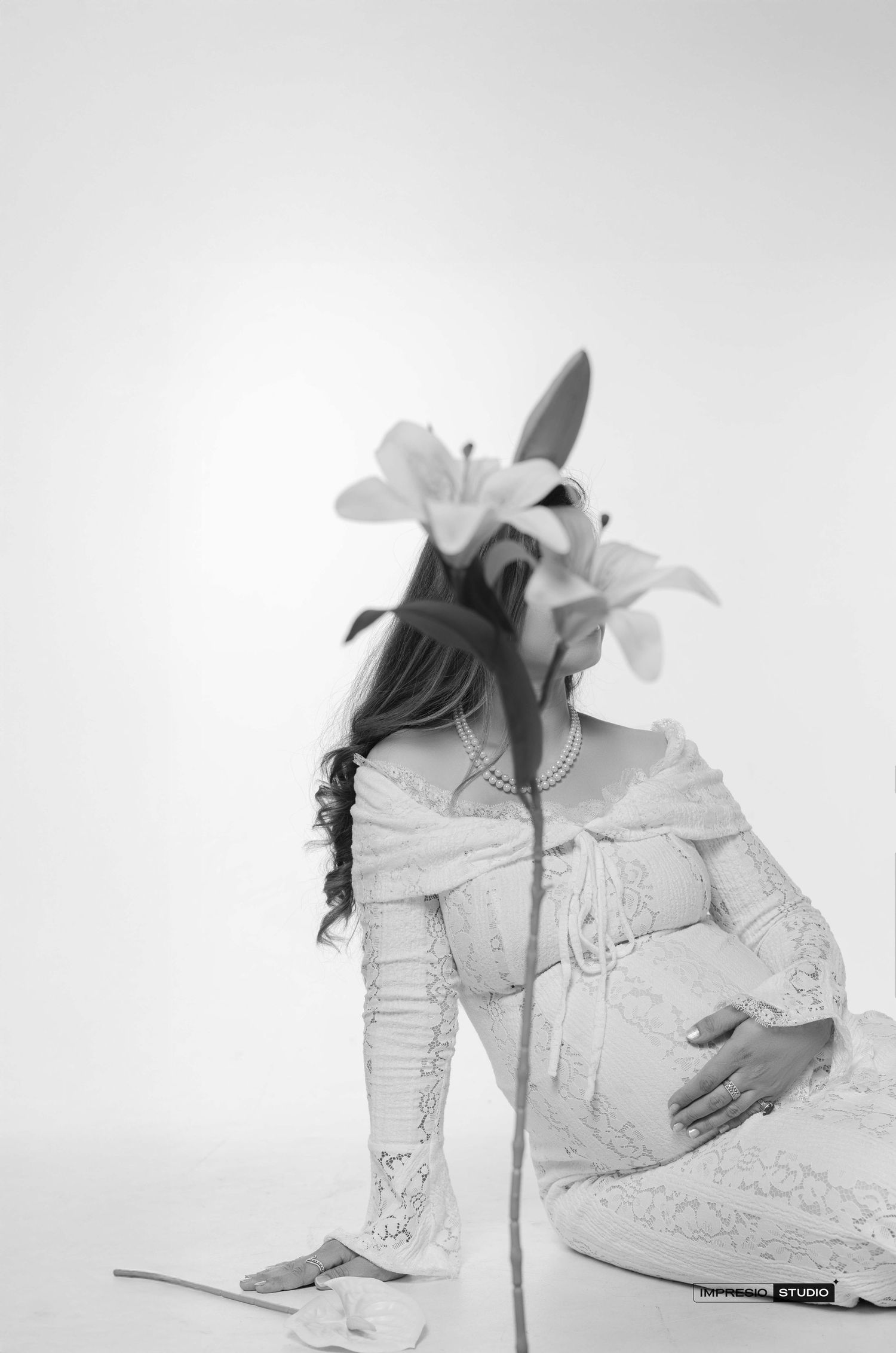 Black and white maternity photo of pregnant woman in lace dress holding belly with lily flower covering her face.